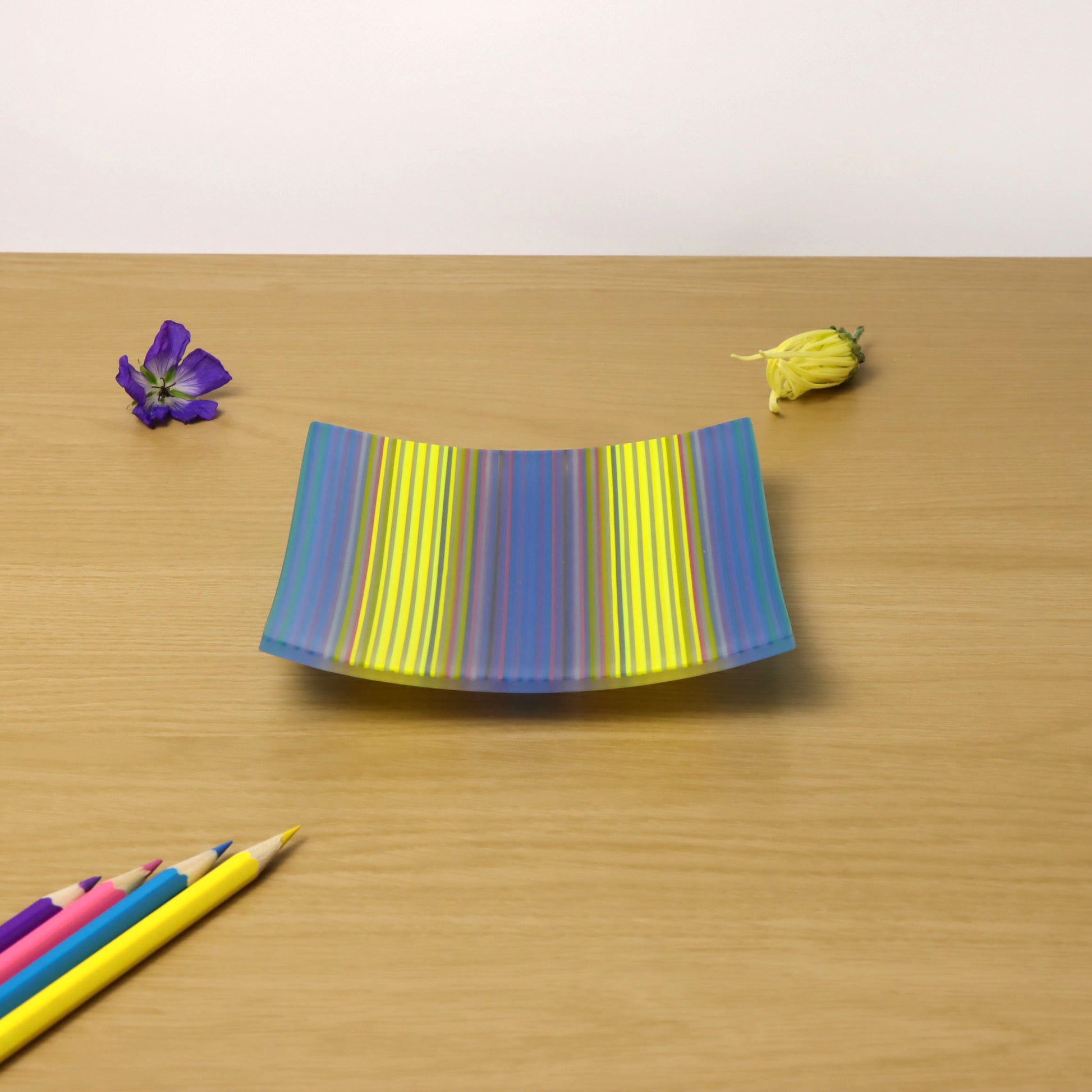 A ColourWave fused glass plate featuring a vibrant spectrum of blue, purple, and yellow vertical stripes is displayed on a wooden surface. To the left of the plate rests a single purple flower with green foliage, and to the right lies a small, unopened yellow bud. In the foreground, a collection of coloured pencils, reflects some of the colours found in the glass plate.