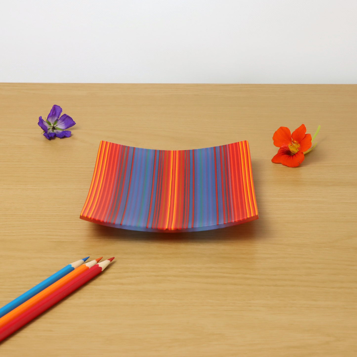 A square ColourWave fused glass plate adorned with vertical stripes in a palette of blue, red, and orange. The plate gently curves at the edges, resting on a wooden surface. Adjacent to the plate are two vibrant flowers, one purple and one orange, each with a yellow centre. In the foreground, a trio of coloured pencils in red, blue, and yellow lie diagonally across the corner, adding a playful touch to the composition.