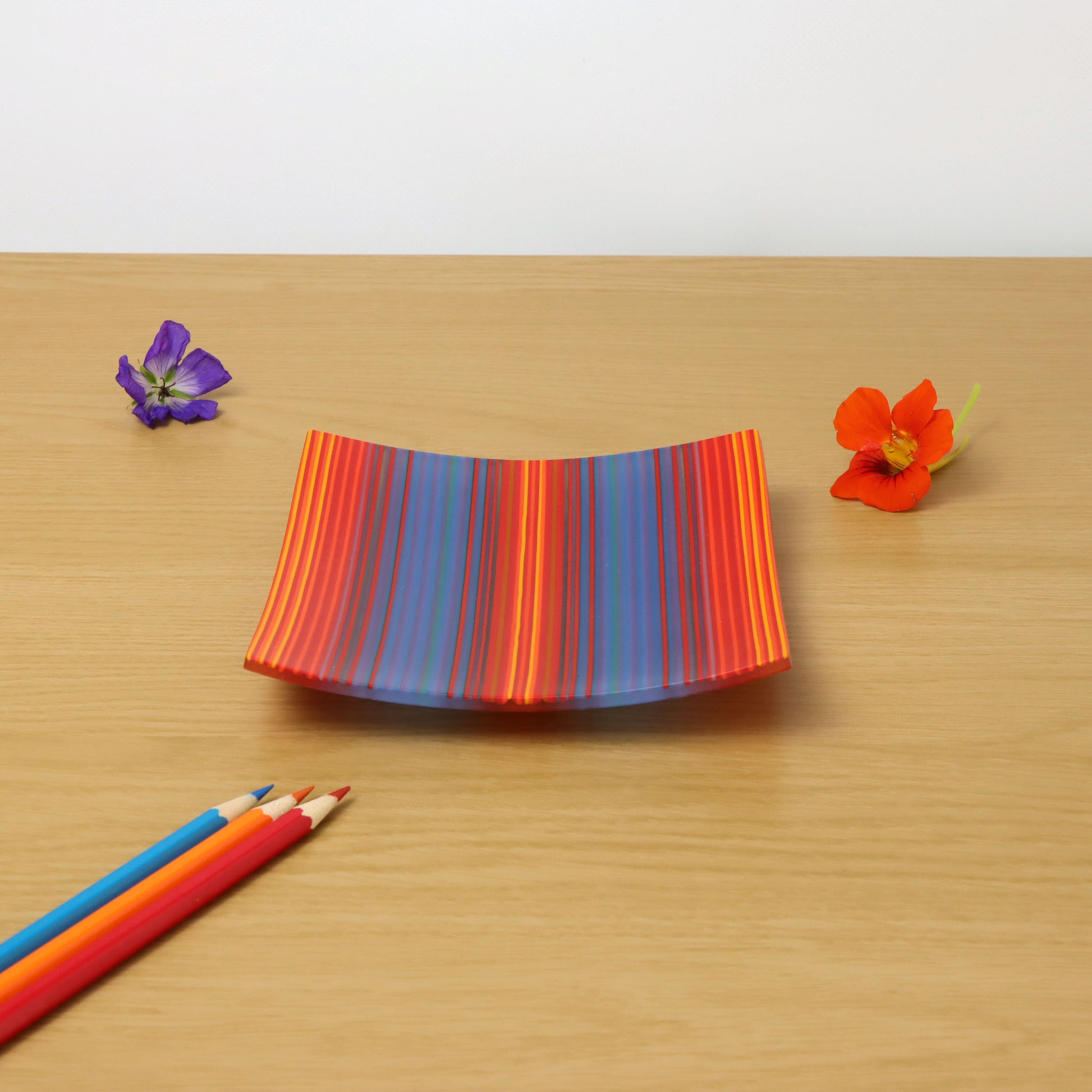 A square ColourWave fused glass plate adorned with vertical stripes in a palette of blue, red, and orange. The plate gently curves at the edges, resting on a wooden surface. Adjacent to the plate are two vibrant flowers, one purple and one orange, each with a yellow centre. In the foreground, a trio of coloured pencils in red, blue, and yellow lie diagonally across the corner, adding a playful touch to the composition.