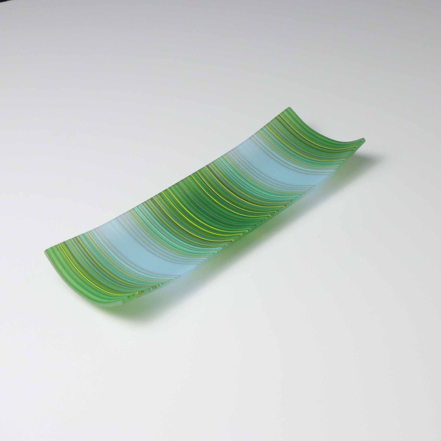 A handcrafted ColourWave Glass fused glass plate, rectangular with corners elegantly curved upwards. It features a harmonious wave pattern in varying shades of green and blue, creating a visual representation of undulating water. The translucent quality of the glass allows light to filter through, accentuating the depth and vibrancy of the colours. The plate exemplifies fine British craftsmanship in fused glass.