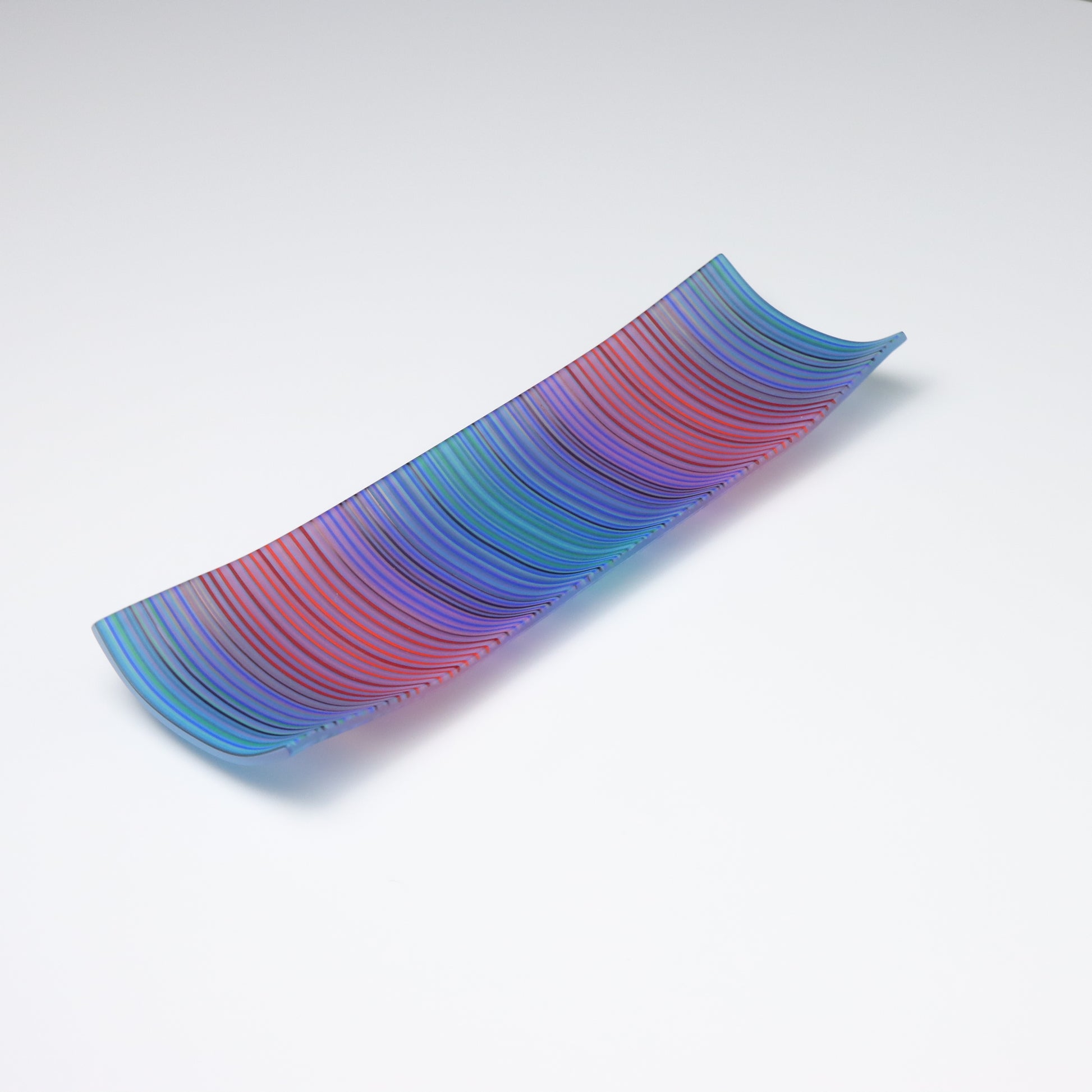 A rectangular ColourWave Glass fused glass plate displaying a vibrant array of vertical stripes in shades of blue, green, red, and purple. The plate exhibits a subtle upward curve at each corner, lending it a sophisticated and contemporary appearance. Its smooth surface catches and reflects the light, showcasing the rich colours and fine strands of glass used to create this piece. Positioned against a white backdrop, the plate’s vivid colours and stylish design are prominently featured.