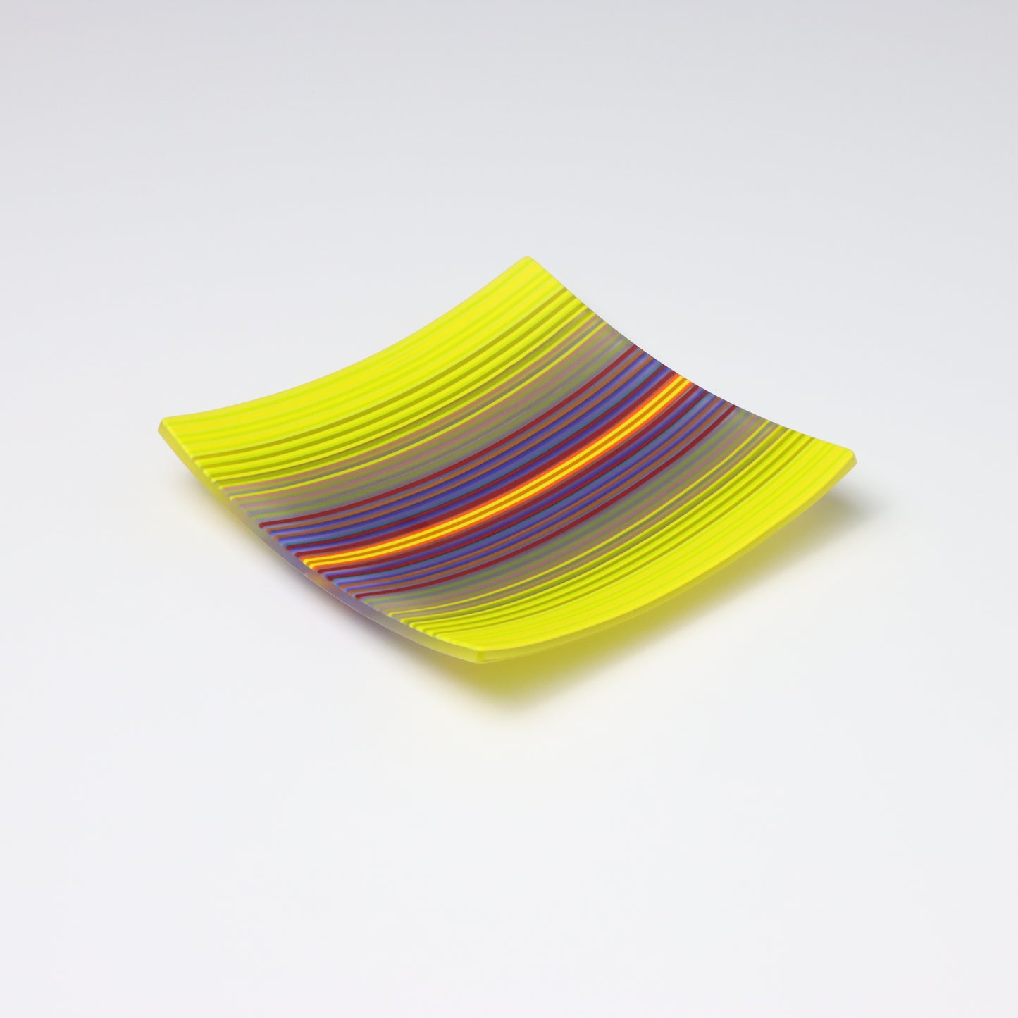 A ColourWave fused glass plate showcasing a mesmerising blend of yellow and purple hues, with strands of red and blue and grey seen up close, adding depth to the dynamic ColourWave pattern. The plate’s square shape and upturned cornerss lend it a contemporary, slightly bowl-like form. The vivid colours a render it a striking piece of decorative art glassware, perfect for adding a splash of colour to any interior space.