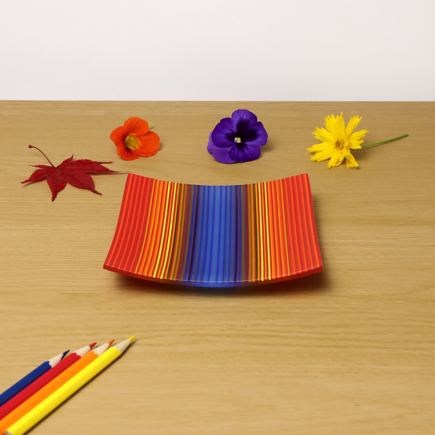 A ColourWave Glass Fused  glass Plate featuring a vibrant array of stripes in shades of red, orange, blue, and yellow. The plate has a square shape with upturned corners and is set against a plain white background. The pattern and the bright colours create an eye-catching visual effect that showcases the intricate craftsmanship of fused glass technique in the UK