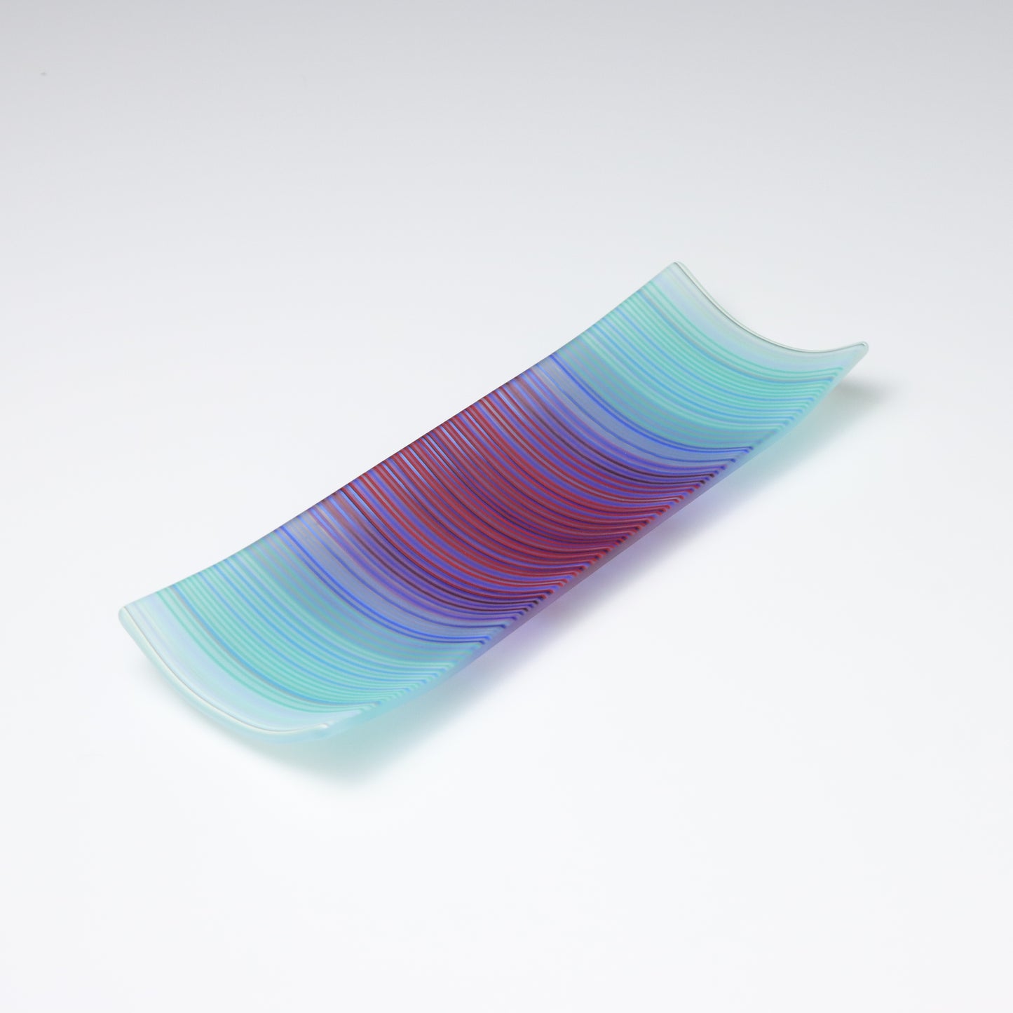 A rectangular ColourWave Glass fused glass plate, boasting a vibrant gradient from turquoise to deep blue and purple in vertical stripes. The plate’s corners elegantly curve upwards, adding a modern twist to its design. The smooth transition of colours offers a serene visual experience, while the craftsmanship reflects a high level of skill in glass work