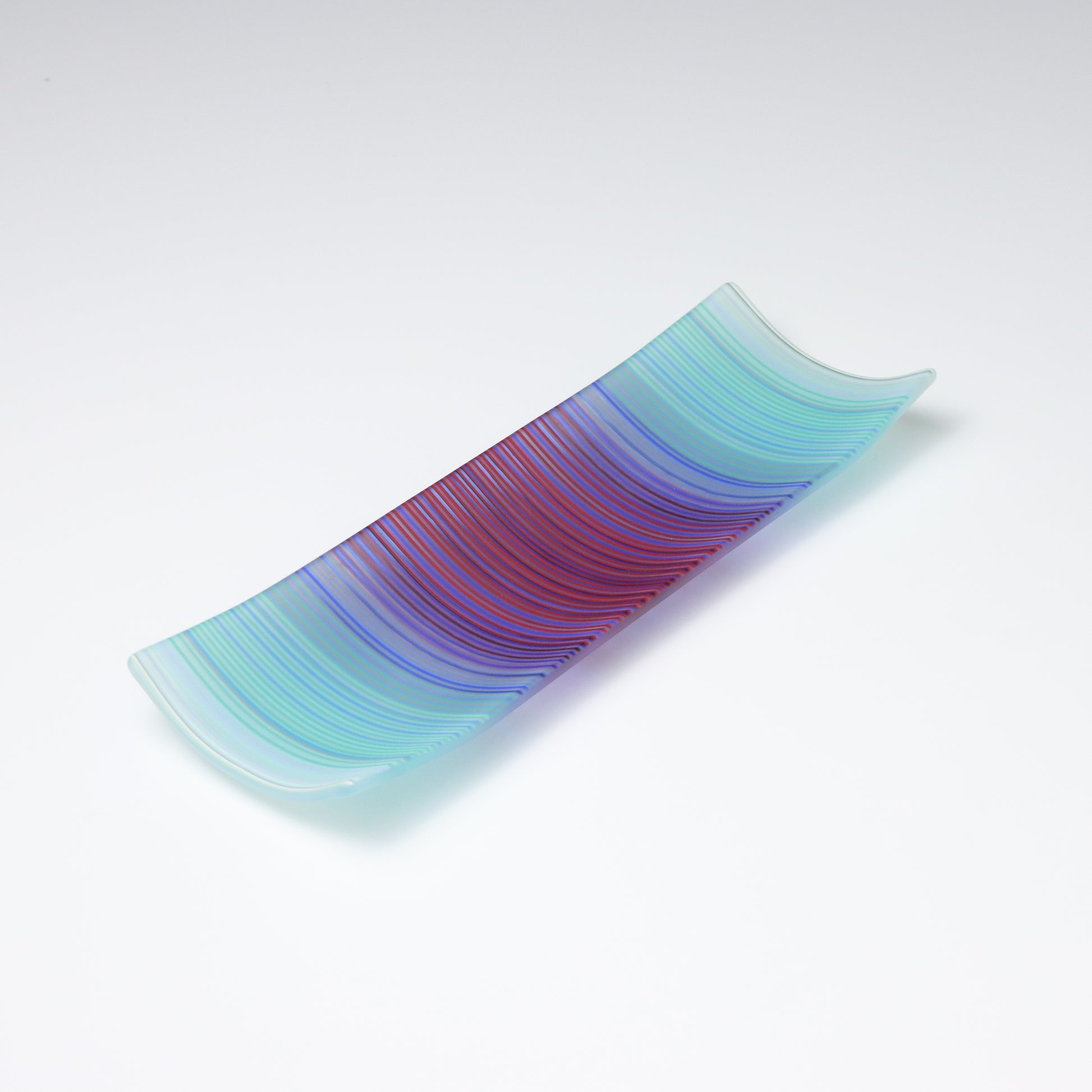 A rectangular ColourWave Glass fused glass plate, boasting a vibrant gradient from turquoise to deep blue and purple in vertical stripes. The plate’s corners elegantly curve upwards, adding a modern twist to its design. The smooth transition of colours offers a serene visual experience, while the craftsmanship reflects a high level of skill in glass work