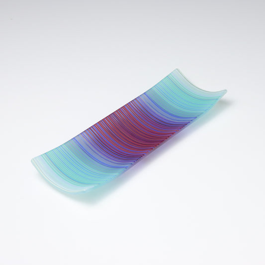 A rectangular ColourWave Glass fused glass plate, boasting a vibrant gradient from turquoise to deep blue and purple in vertical stripes. The plate’s corners elegantly curve upwards, adding a modern twist to its design. The smooth transition of colours offers a serene visual experience, while the craftsmanship reflects a high level of skill in glass work