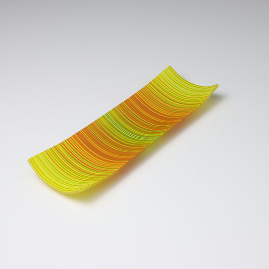 A ColourWave Glass fused glass plate, adorned with a vibrant pattern of vertical stripes in shades of yellow, green, orange, and red. The rectangular plate gently curves at the corners, forming a shallow bowl shape. The bright colours and sleek design emphasize its role as an attractive decorative piece.