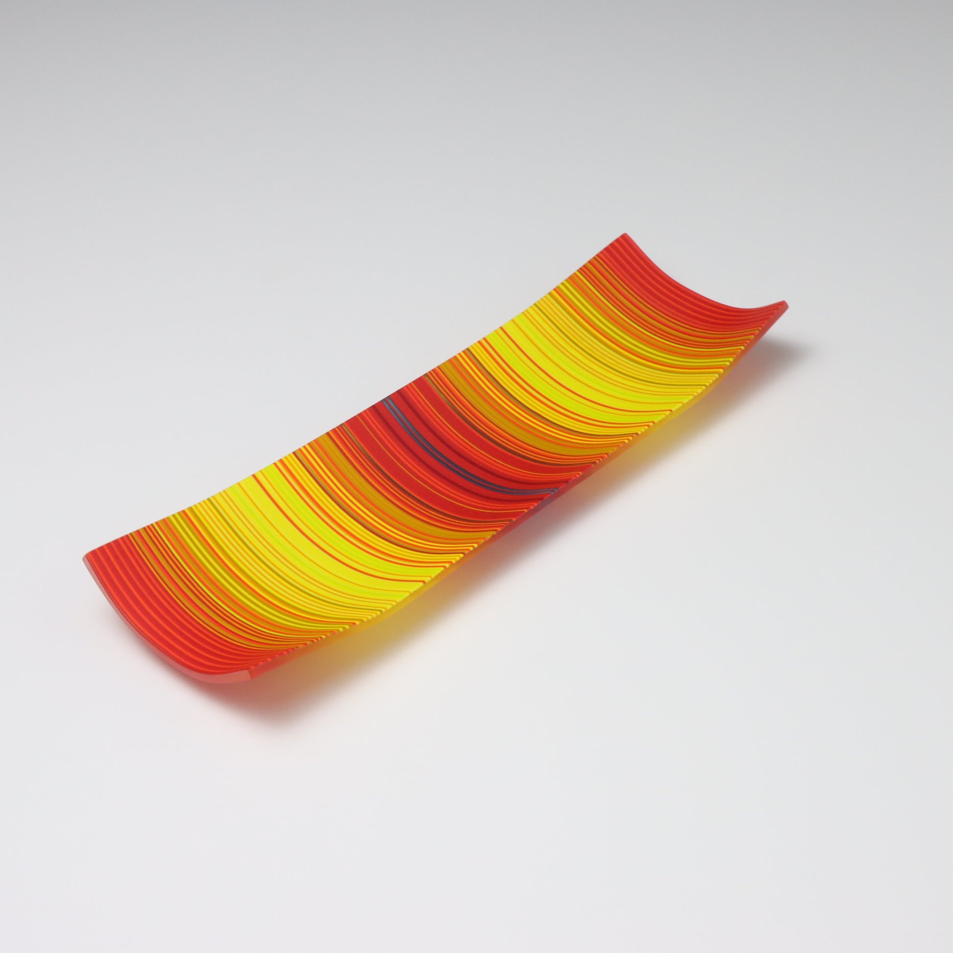 A captivating ColourWave Glass fused glass plate, showcasing a vibrant colour gradient that transitions between bright red to bright yellow. The plate’s rectangular shape is softened by the corners that curve gently upwards, adding an elegant touch to the design. Set against a neutral grey backdrop, the plate’s rich colours and sleek form are beautifully highlighted.
