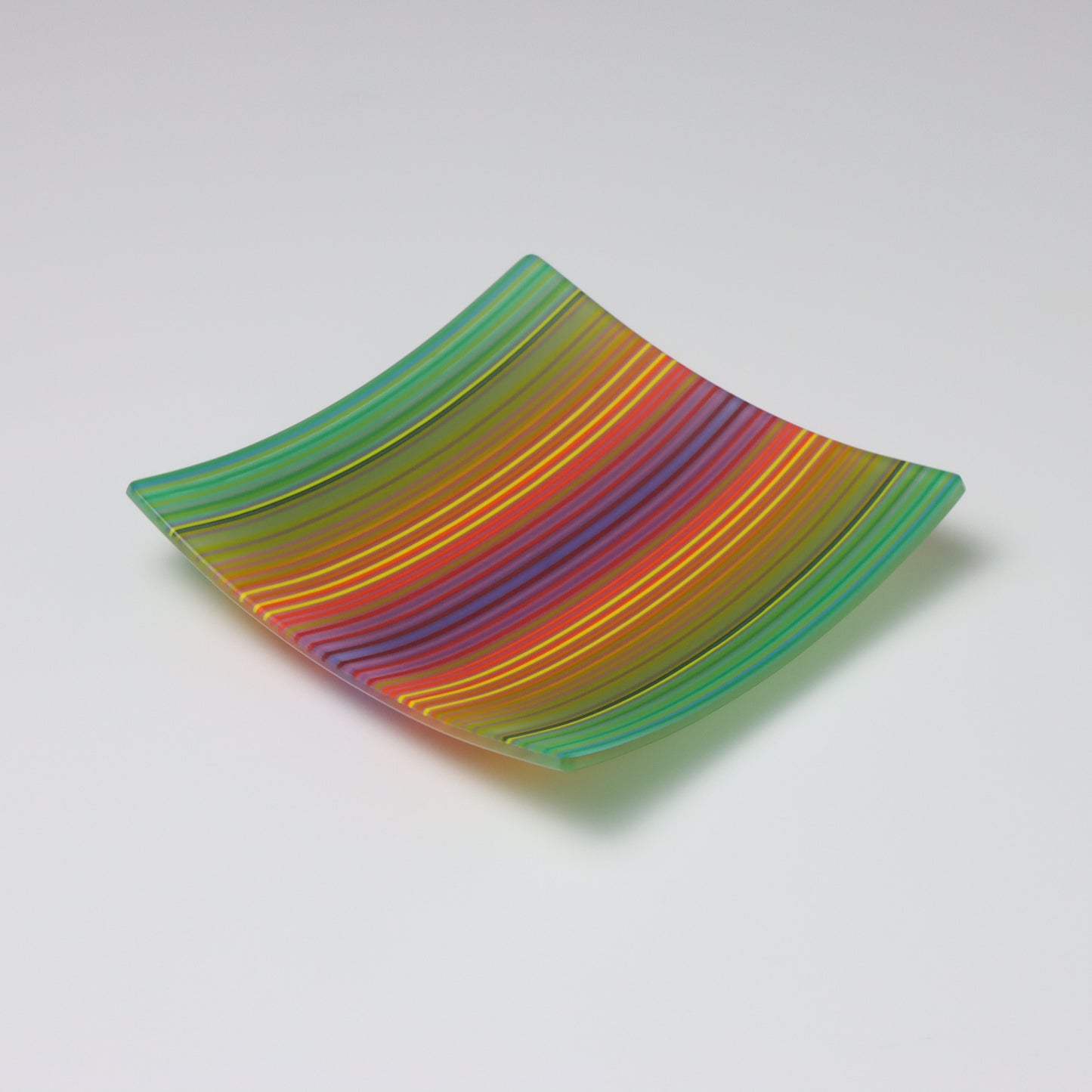 A ColourWave Glass fused glass plate with a vibrant spectrum of parallel stripes in shades of green, yellow, orange, and red, with many more when seen close up. The square-shaped plate has gently upturned corners, forming a shallow bowl. The smooth transitions between colours create a dynamic, wave-like effect, showcasing the intricate skill of fused glass artistry.