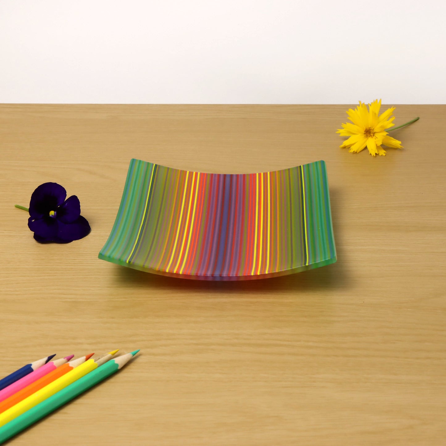 A ColourWave Glass fused glass plate featuring vibrant vertical stripes in a spectrum of colours including green, blue, yellow, red, and purple. The square plate is slightly curved upwards at the corners and rests on a wooden surface. Adjacent to the plate, a deep purple pansy and a bright yellow daisy add a touch of natural beauty, while a set of coloured pencils in the foreground suggests an artistic setting.