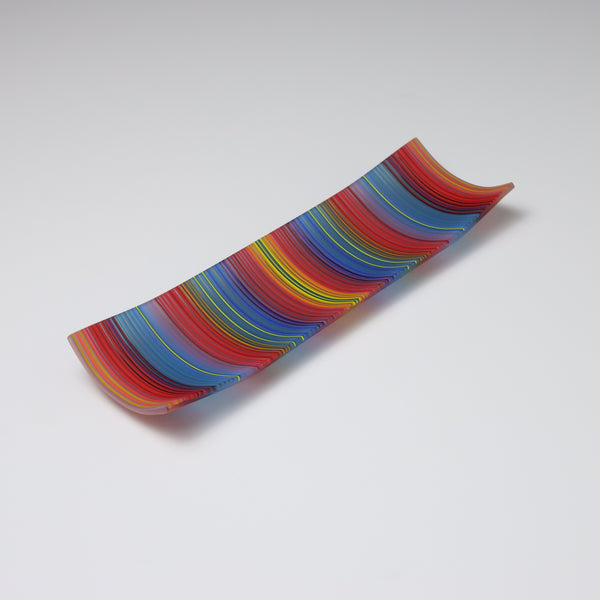 A ColourWave Glass fused glass plate, arrayed with vertical stripes in vibrant red, blue, yellow, and green. The plate’s rectangular shape is enhanced by the corners that curve upward, forming a shallow, dish-like appearance. Set against a grey background, the plate’s rich stripes and distinctive design are highlighted, making it an eye-catching piece.