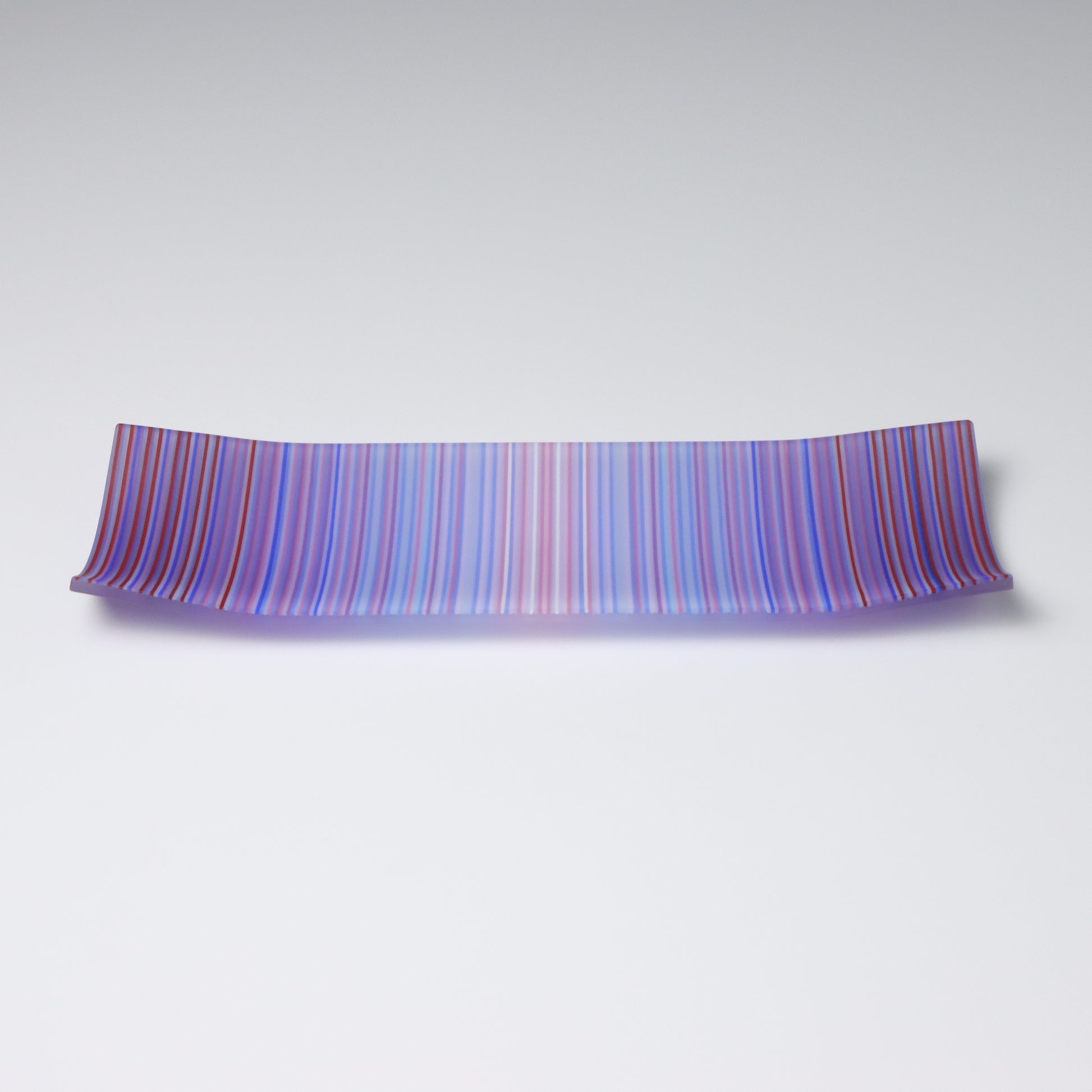 A rectangular fused glass plate showcasing a ColourWave design. The design features a captivating blend of purple, blue, and subtle pink stripes. The surface of the plate is smooth, reflecting light and highlighting the rich colours and the meticulous craftsmanship that went into its creation. The plate is set against a neutral grey gradient background, which further accentuates its vibrant hues and elegant form.