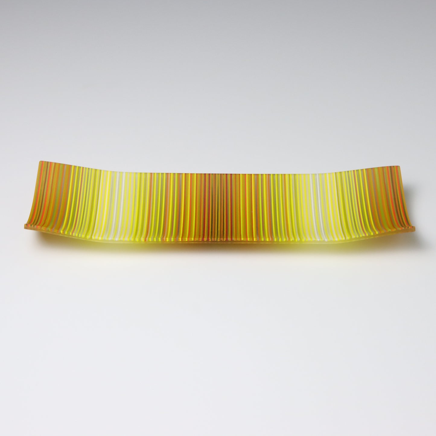 A rectangular ColourWave fused glass plate, boasting vibrant vertical stripes in shades of yellow, orange, and green. The plate’s design features a gentle upward curve at each corner, giving it a subtle, shallow vessel shape suitable for artistic display.