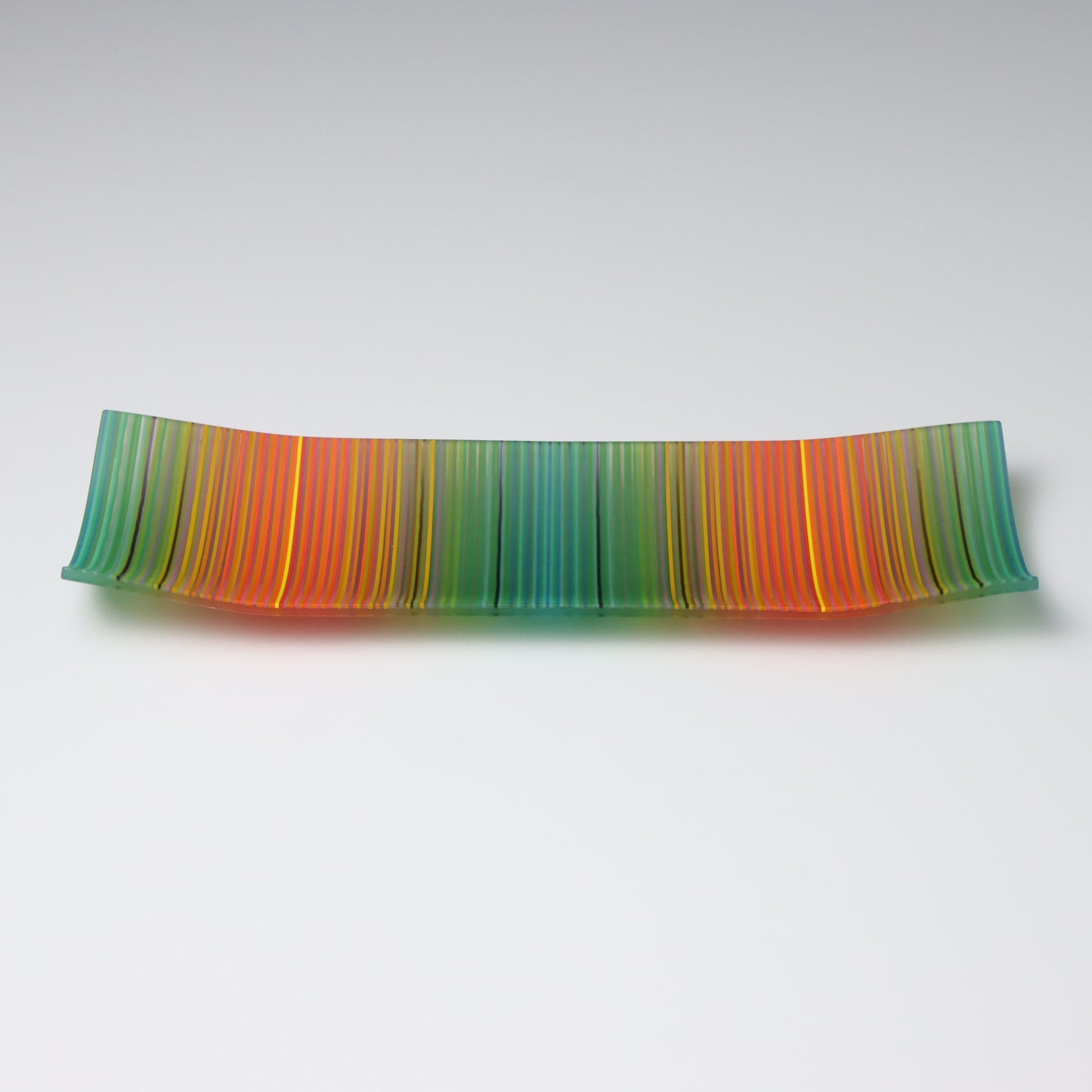 A vibrant ColourWave Glass fused glass plate, boasting a striking striped pattern with a gradient of greens, oranges, and reds. The rectangular plate features elegantly upturned corners, creating a shallow, bowl-like form that exudes sophistication.