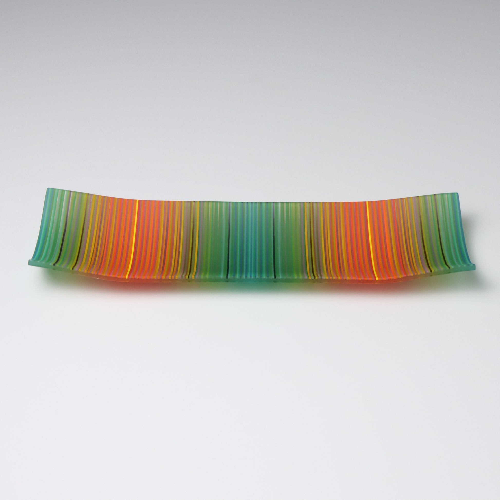 A vibrant ColourWave Glass fused glass plate, boasting a striking striped pattern with a gradient of greens, oranges, and reds. The rectangular plate features elegantly upturned corners, creating a shallow, bowl-like form that exudes sophistication.