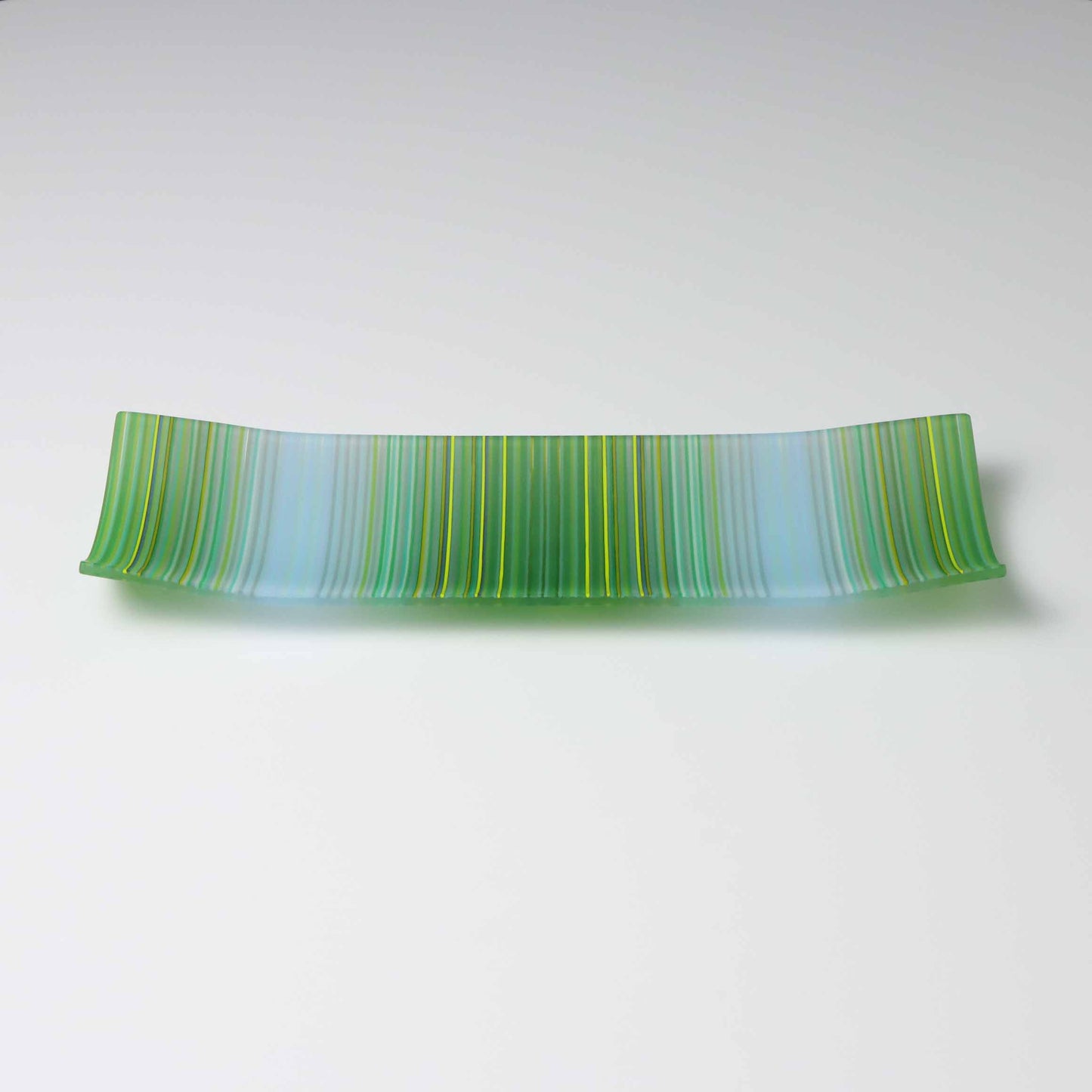 A handcrafted ColourWave Glass fused glass plate, rectangular with corners elegantly curved upwards. It features a harmonious wave pattern in varying shades of green and blue, creating a visual representation of undulating water. The translucent quality of the glass allows light to filter through, accentuating the depth and vibrancy of the colours. The plate exemplifies fine British craftsmanship in fused glass.