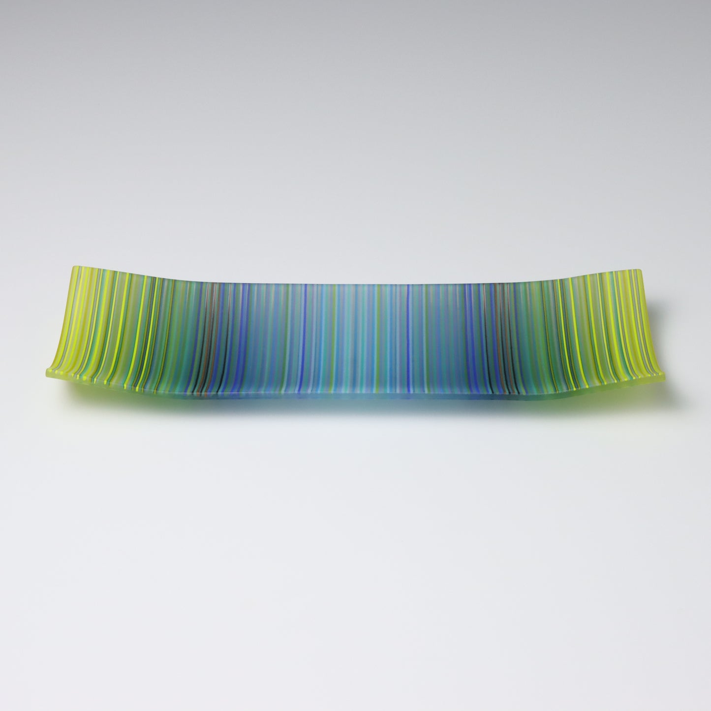 A rectangular ColourWave Glass plate, artistically crafted with a series of vertical stripes cascading through shades of blue and green. The plate’s corners rise subtly, giving it a unique and decorative shape. This piece is a testament to contemporary design, enhancing the visual appeal of any room with its artistic presence.