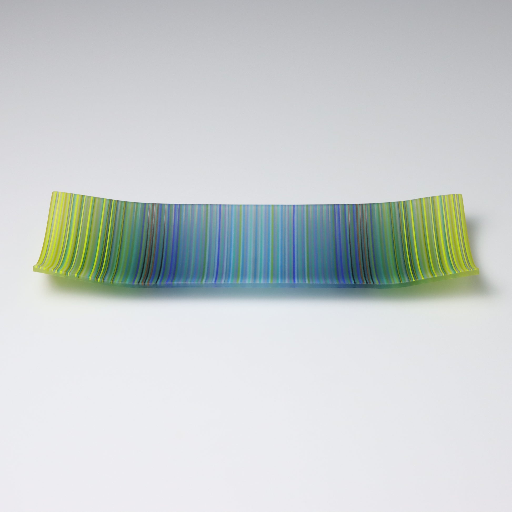 A rectangular ColourWave Glass plate, artistically crafted with a series of vertical stripes cascading through shades of blue and green. The plate’s corners rise subtly, giving it a unique and decorative shape. This piece is a testament to contemporary design, enhancing the visual appeal of any room with its artistic presence.