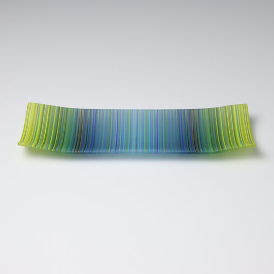 A rectangular ColourWave Glass plate, artistically crafted with a series of vertical stripes cascading through shades of blue and green. The plate’s corners rise subtly, giving it a unique and decorative shape. This piece is a testament to contemporary design, enhancing the visual appeal of any room with its artistic presence.