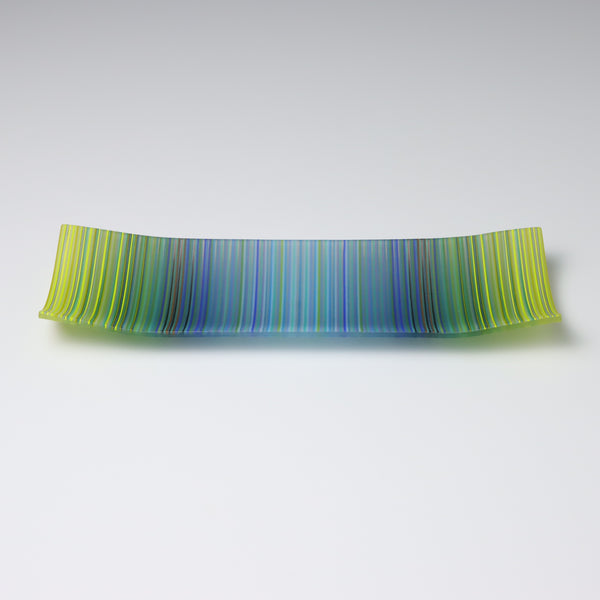 A rectangular ColourWave Glass plate, artistically crafted with a series of vertical stripes cascading through shades of blue and green. The plate’s corners rise subtly, giving it a unique and decorative shape. This piece is a testament to contemporary design, enhancing the visual appeal of any room with its artistic presence.