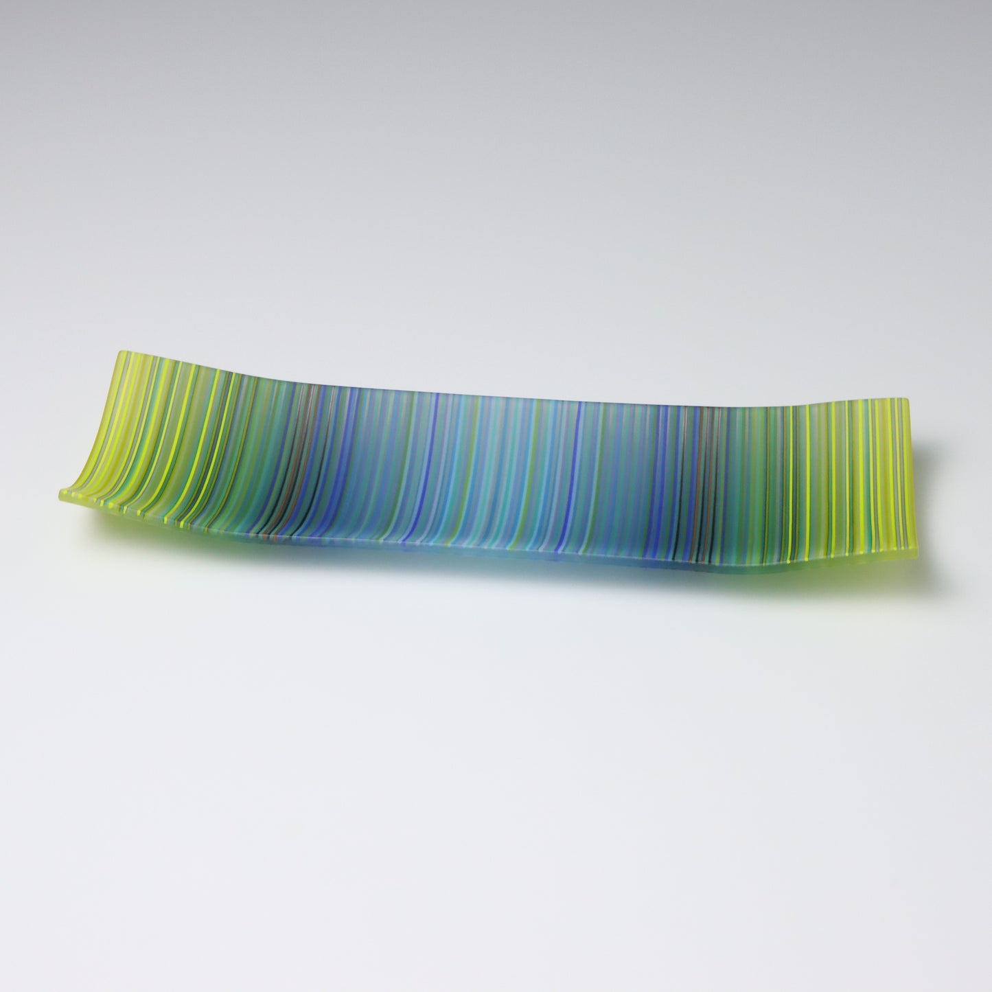 A rectangular ColourWave Glass plate, artistically crafted with a series of vertical stripes cascading through shades of blue and green. The plate’s corners rise subtly, giving it a unique and decorative shape. This piece is a testament to contemporary design, enhancing the visual appeal of any room with its artistic presence.