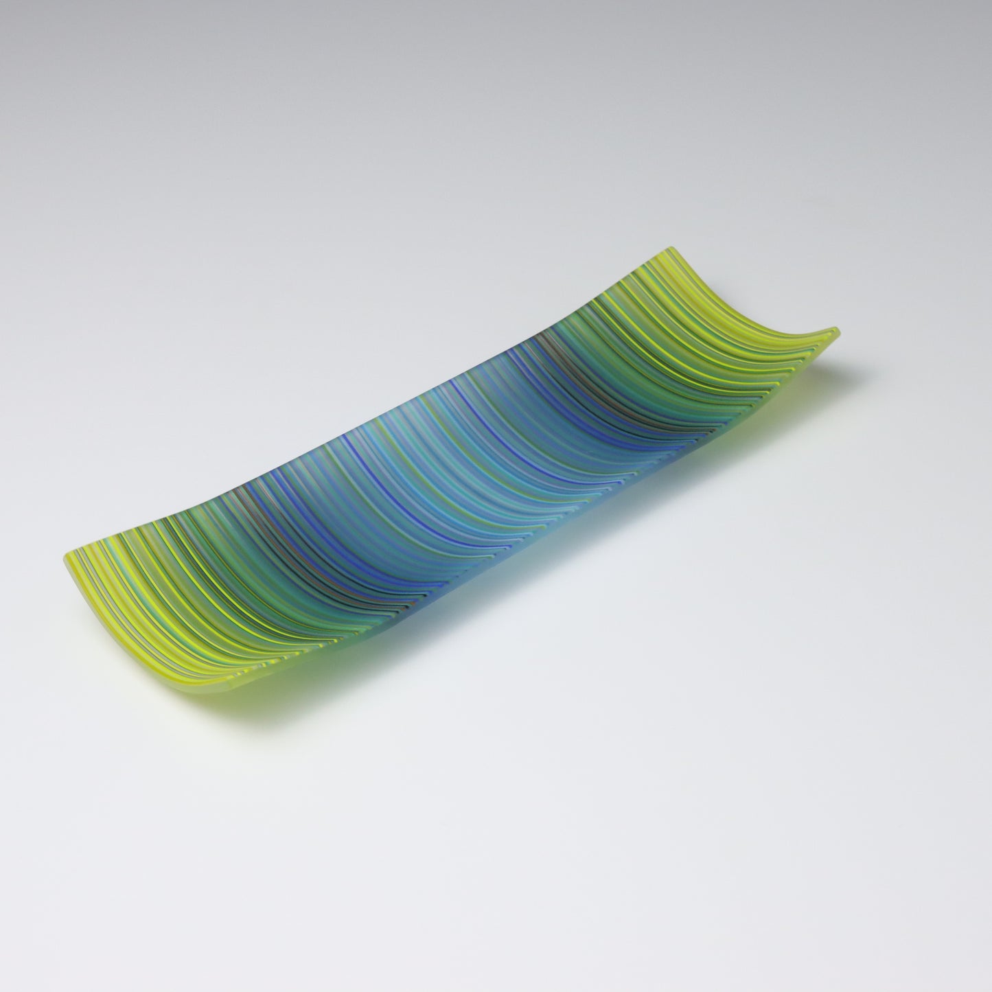 A rectangular ColourWave Glass plate, artistically crafted with a series of vertical stripes cascading through shades of blue and green. The plate’s corners rise subtly, giving it a unique and decorative shape. This piece is a testament to contemporary design, enhancing the visual appeal of any room with its artistic presence.