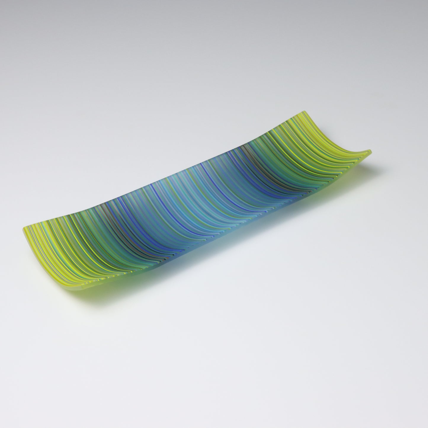 A rectangular ColourWave Glass plate, artistically crafted with a series of vertical stripes cascading through shades of blue and green. The plate’s corners rise subtly, giving it a unique and decorative shape. This piece is a testament to contemporary design, enhancing the visual appeal of any room with its artistic presence.
