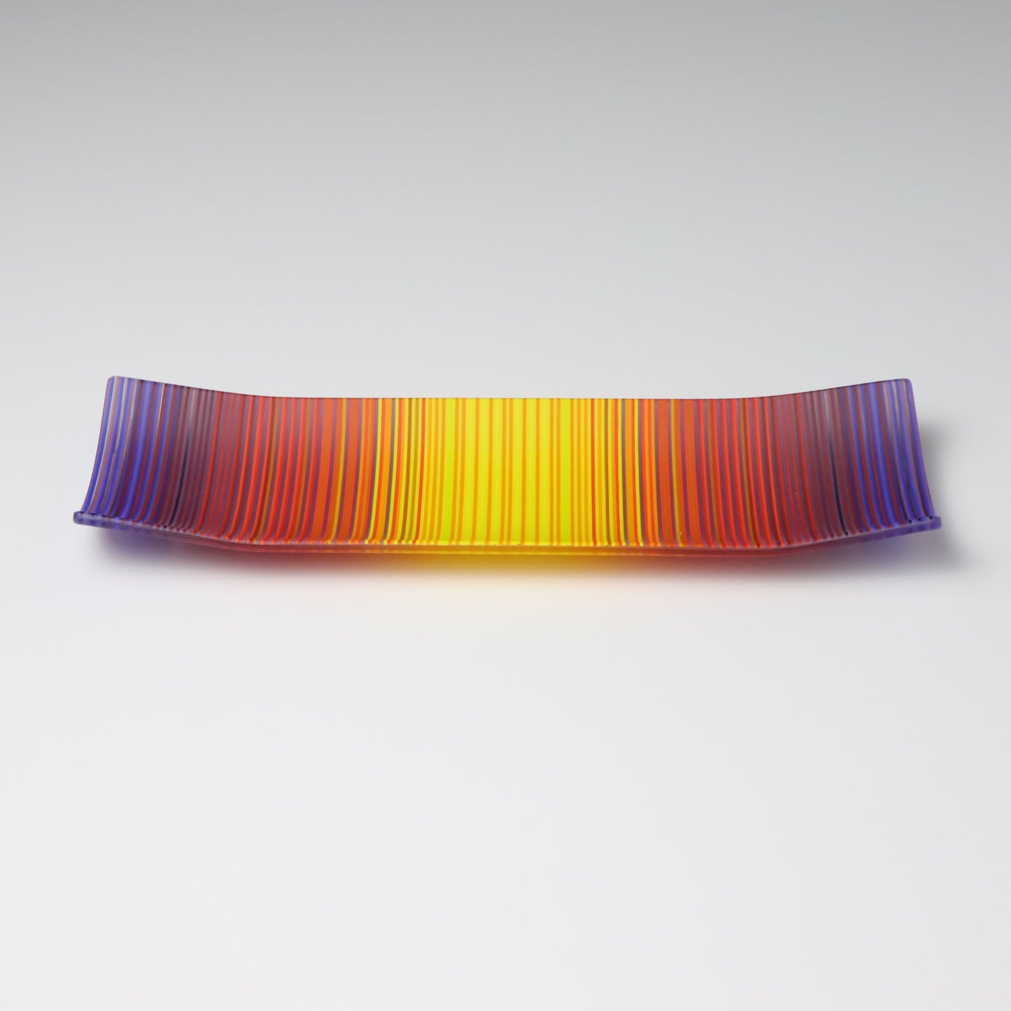 A rectangular ColourWave Glass fused glass plate showcasing a vivid spectrum of colours, with purple at the edges blending into red, orange, and yellow towards the centre. The plate has a smooth surface and its corners gently curve upwards, giving it a contemporary and elegant profile.