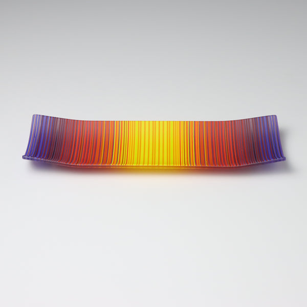 A rectangular ColourWave Glass fused glass plate showcasing a vivid spectrum of colours, with purple at the edges blending into red, orange, and yellow towards the centre. The plate has a smooth surface and its corners gently curve upwards, giving it a contemporary and elegant profile.