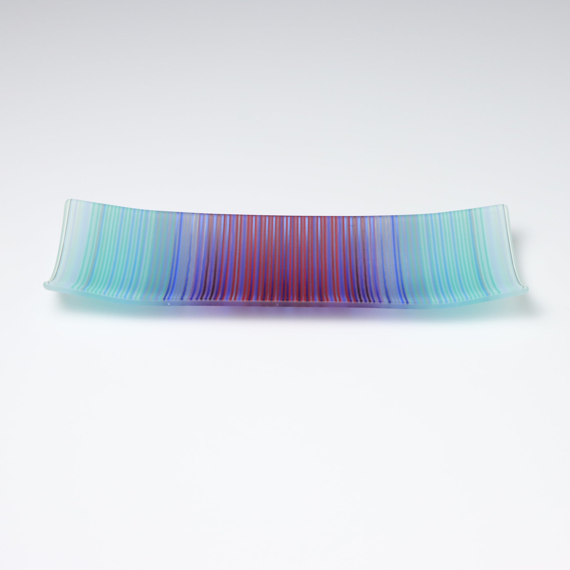 A rectangular ColourWave Glass fused glass plate, boasting a vibrant gradient from turquoise to deep blue and purple in vertical stripes. The plate’s corners elegantly curve upwards, adding a modern twist to its design. The smooth transition of colours offers a serene visual experience, while the craftsmanship reflects a high level of skill in glass work