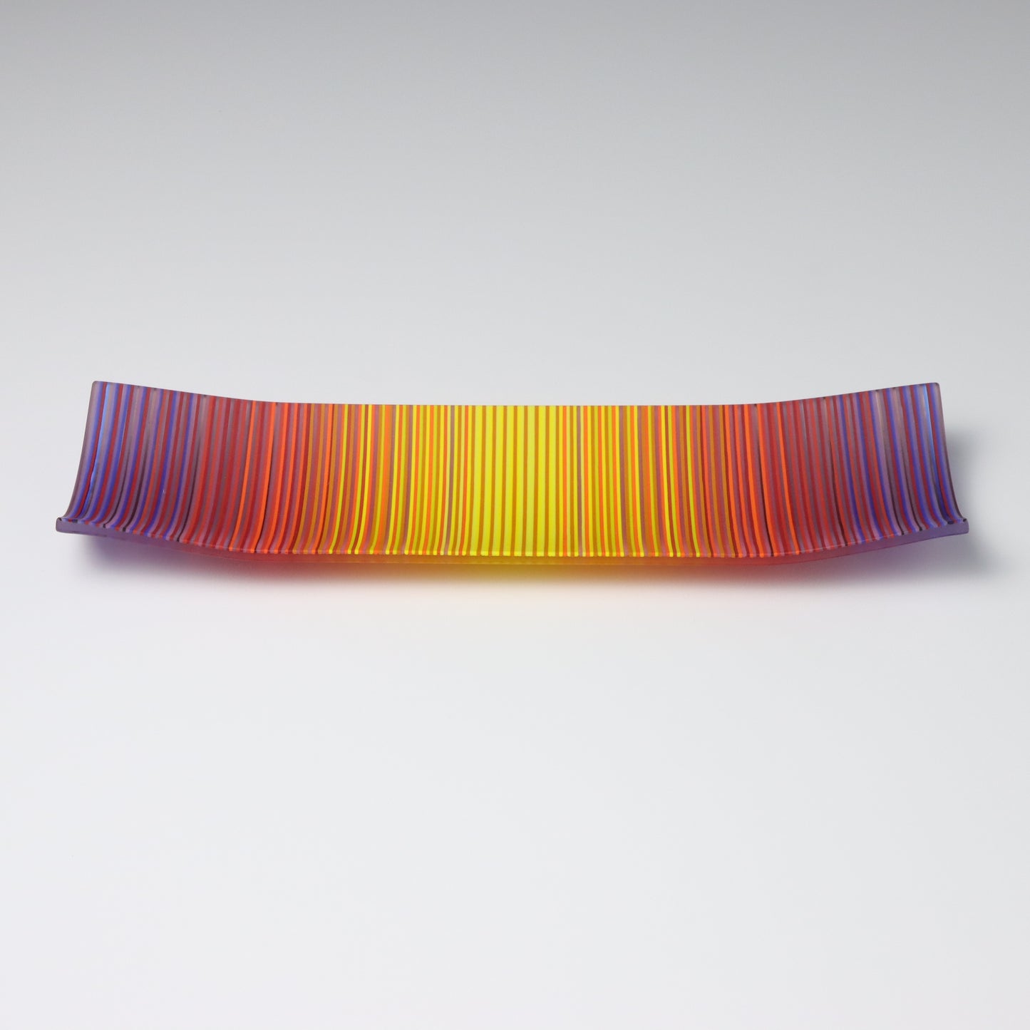 A ColourWave Glass fused glass plate, rectangular in shape, displays vibrant stripes in shades of purple, red, orange, and yellow. The plate is distinctively curved upwards at each corner, forming an elegant raised edge. It rests on a wooden surface, complemented by a colourful arrangement of flowers and a set of pencils that match the plate’s vivid stripes.