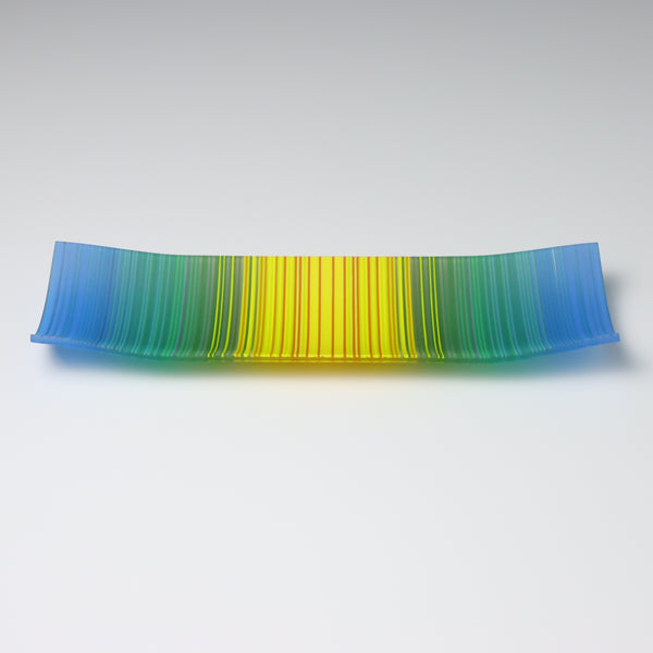 A ColourWave Glass fused glass plate displaying a vibrant gradient, with hues transitioning from blue at the edges to green and culminating in yellow at the centre. The plate’s rectangular form is accentuated by its elegantly upturned corners, lending a modern and sophisticated touch to its design.