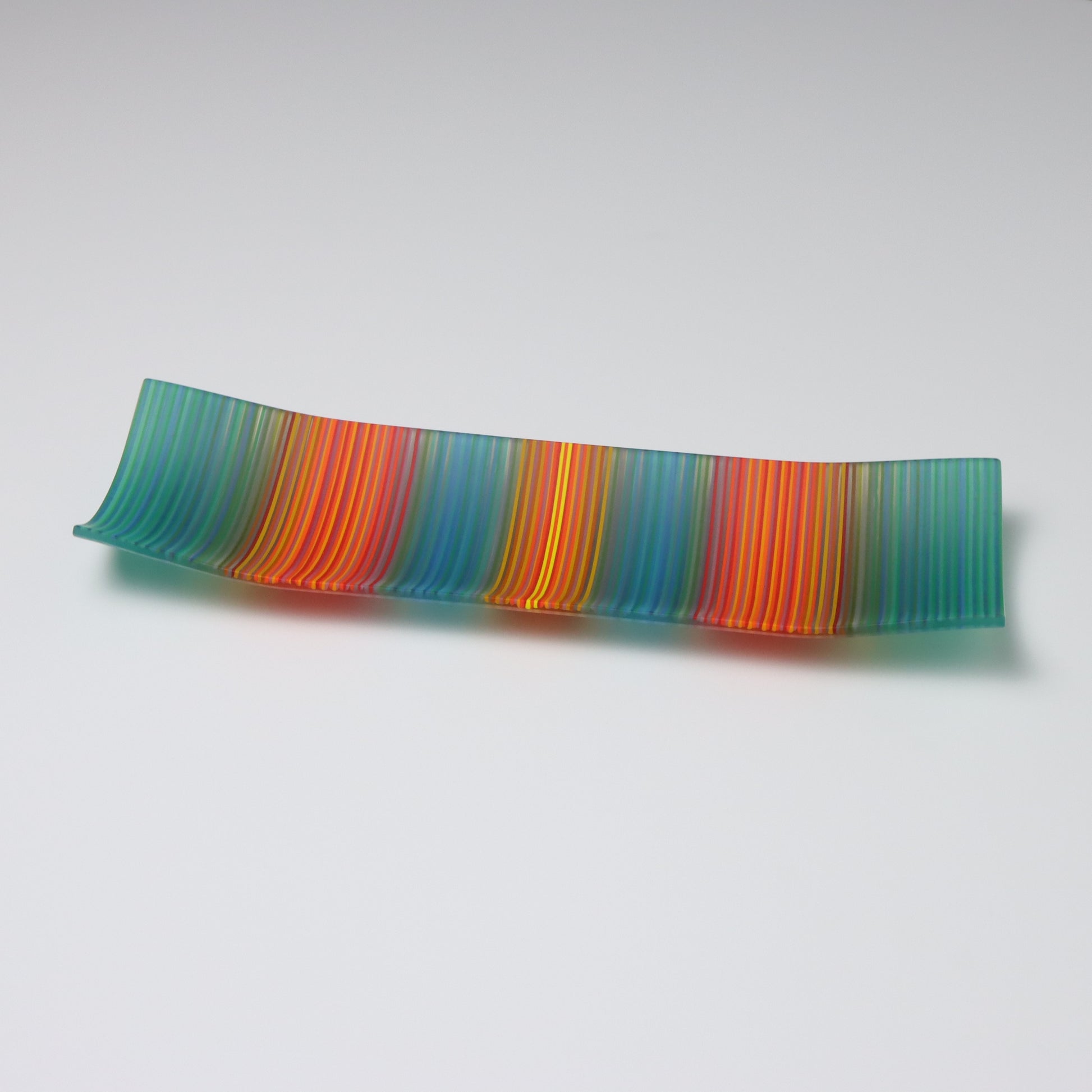 Displayed against a white backdrop is a rectangular fused glass plate from ColourWave Glass. This handcrafted piece features vertical stripes in a soothing teal green, tranquil blue, fiery red, and vibrant orange, with a radiant yellow highlight at the center. The plate’s unique design includes gentle upward curves at the corners and sides, adding a three-dimensional aspect to its visual appeal.