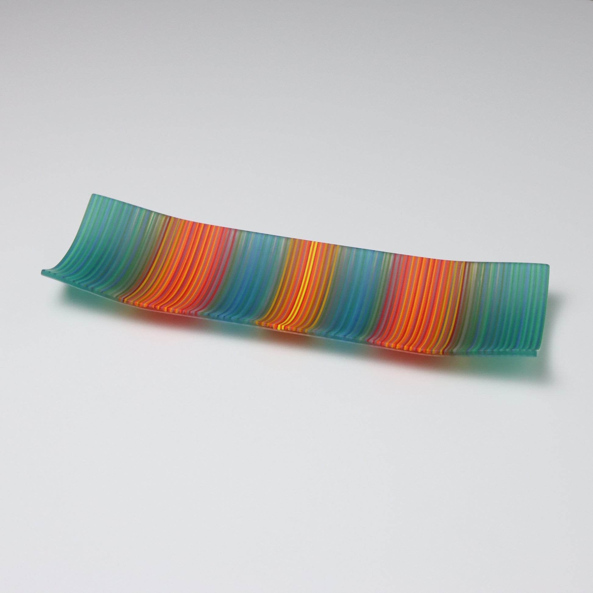 Displayed against a white backdrop is a rectangular fused glass plate from ColourWave Glass. This handcrafted piece features vertical stripes in a soothing teal green, tranquil blue, fiery red, and vibrant orange, with a radiant yellow highlight at the center. The plate’s unique design includes gentle upward curves at the corners and sides, adding a three-dimensional aspect to its visual appeal.