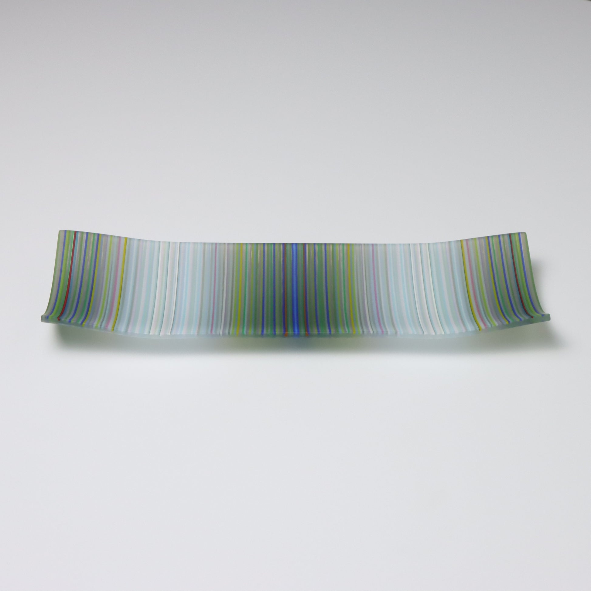 A handcrafted and kilnfired ColourWave Glass plate, rectangular with upturned corners creating a shallow bowl effect. The surface displays a beautiful wave pattern predominantly in grey and white, which upon closer inspection, is composed of a complex mix of many colours. The plate is set against a white background, emphasising its vibrant colours and artistic flair.