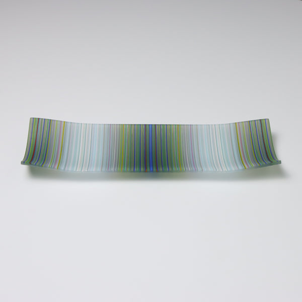 A handcrafted and kilnfired ColourWave Glass plate, rectangular with upturned corners creating a shallow bowl effect. The surface displays a beautiful wave pattern predominantly in grey and white, which upon closer inspection, is composed of a complex mix of many colours. The plate is set against a white background, emphasising its vibrant colours and artistic flair.
