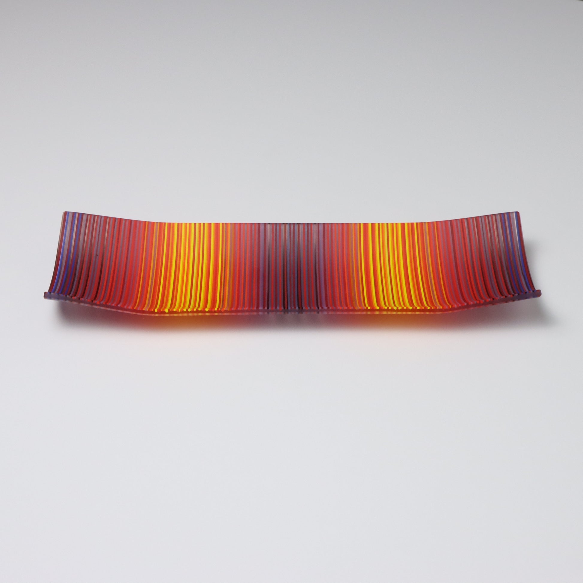 A rectangular ColourWave Glass fused glass plate, with a dynamic gradient of warm colours transitioning from deep reds to warm sunny yellows. The plate’s longer sides elegantly curve upwards at the corners, adding a modern twist to the classic design. The striated pattern across the surface gives the impression of a vivid sunset, making it a striking centrepiece for that special space in your home.