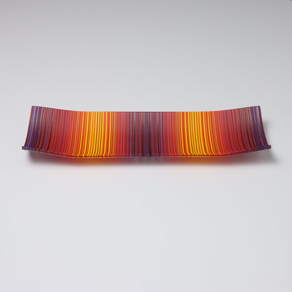 A rectangular ColourWave Glass fused glass plate, with a dynamic gradient of warm colours transitioning from deep reds to warm sunny yellows. The plate’s longer sides elegantly curve upwards at the corners, adding a modern twist to the classic design. The striated pattern across the surface gives the impression of a vivid sunset, making it a striking centrepiece for that special space in your home.
