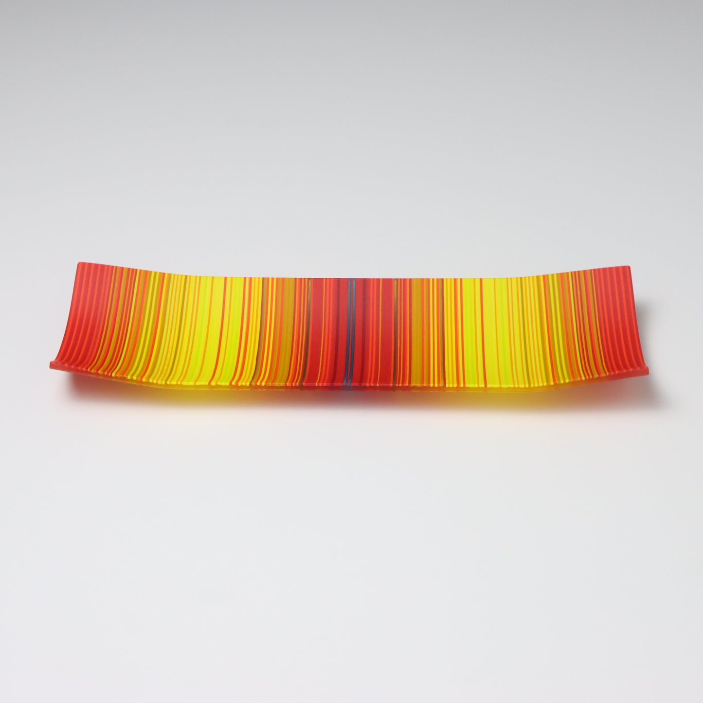 A captivating ColourWave Glass fused glass plate, showcasing a vibrant colour gradient that transitions between bright red to bright yellow. The plate’s rectangular shape is softened by the corners that curve gently upwards, adding an elegant touch to the design. Set against a neutral grey backdrop, the plate’s rich colours and sleek form are beautifully highlighted.