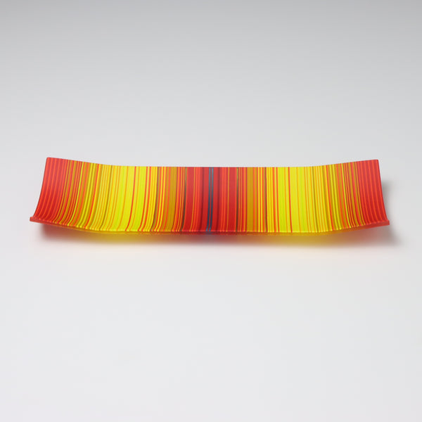 A captivating ColourWave Glass fused glass plate, showcasing a vibrant colour gradient that transitions between bright red to bright yellow. The plate’s rectangular shape is softened by the corners that curve gently upwards, adding an elegant touch to the design. Set against a neutral grey backdrop, the plate’s rich colours and sleek form are beautifully highlighted.