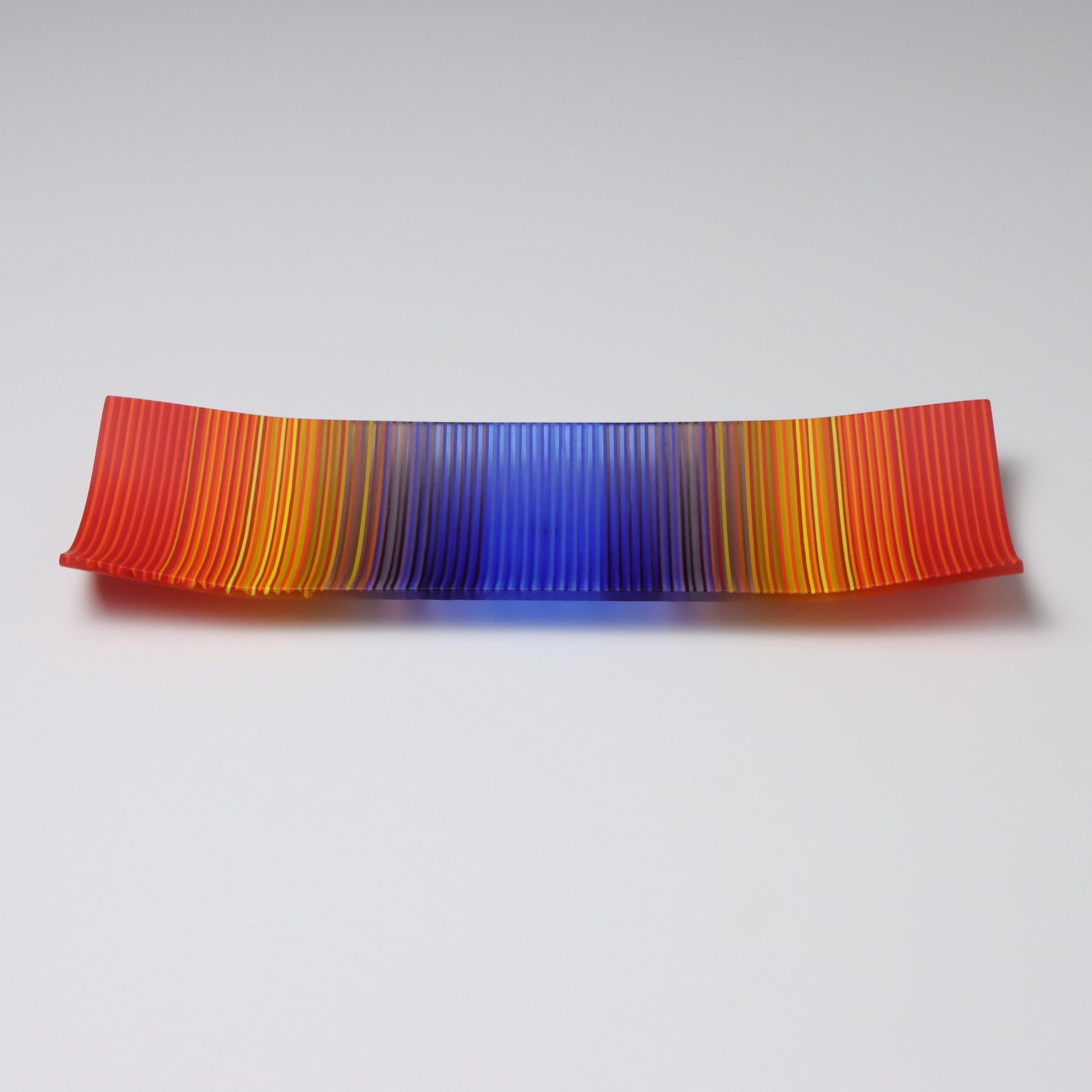 A ColourWave Glass fused glass plate with an eye-catching design. The plate is rectangular with corners that curve upwards. The colour scheme is a vibrant gradient that transitions from red at the edges to deep yellow, then purple, and culminates in a bright blue centre. The smooth blending of colours gives the plate a dynamic and visually striking appearance. It’s a perfect addition to any setting.