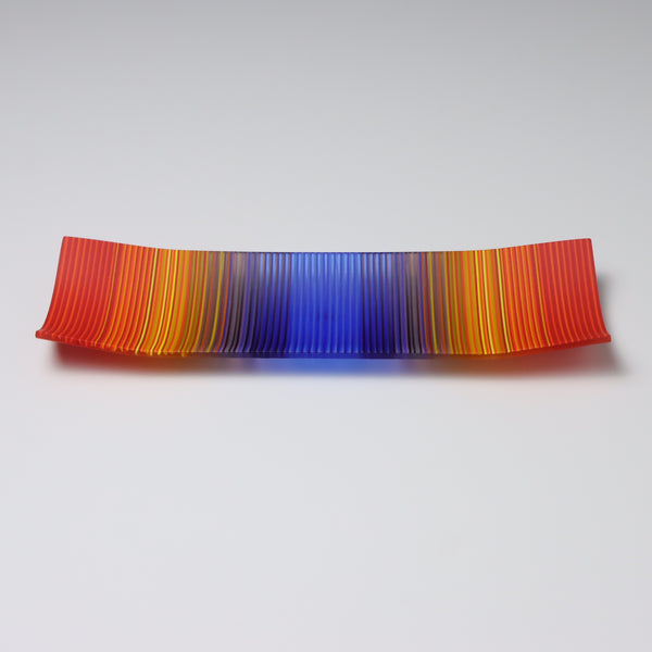 A ColourWave Glass fused glass plate with an eye-catching design. The plate is rectangular with corners that curve upwards. The colour scheme is a vibrant gradient that transitions from red at the edges to deep yellow, then purple, and culminates in a bright blue centre. The smooth blending of colours gives the plate a dynamic and visually striking appearance. It’s a perfect addition to any setting.