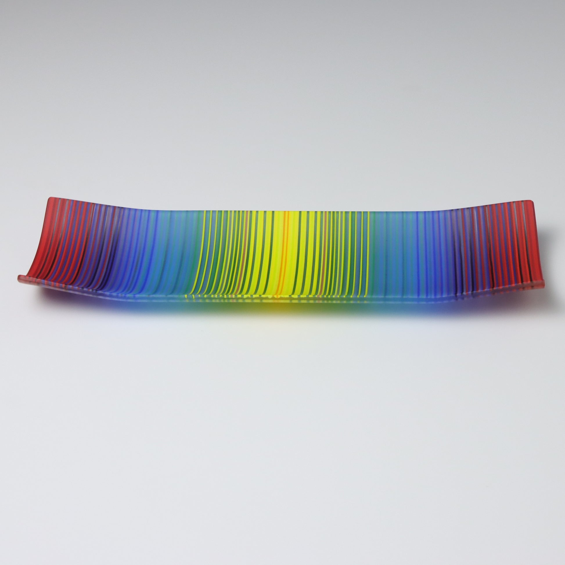 A ColourWave Glass fused glass plate, showcasing a vibrant spectrum of colours that transition smoothly from one to another, reminiscent of a rainbow just the colours are not in the same order!. The plate’s design features gracefully curving corners that add a touch of elegance to its rectangular shape. It is placed on a plain white background letting the bright colours of this decorative fused glass plate shine.
