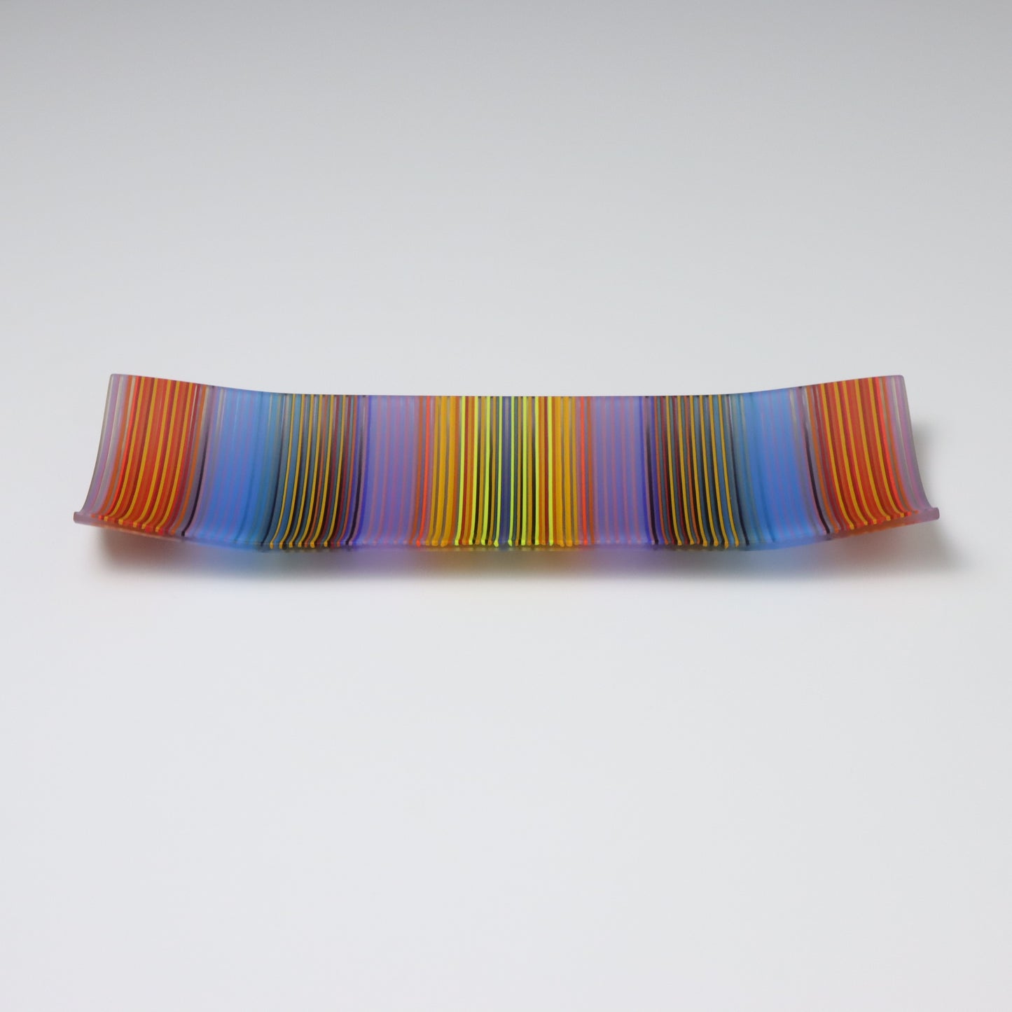 An exquisite ColourWave Glass plate with a rectangular shape, featuring a striking pattern of vertical stripes in a bold colour palette including shades of blue, purple, orange, yellow and green, with many more seen up close. The design is complemented by the plate’s corners, which gracefully curve upwards, giving it a refined and elevated appearance.