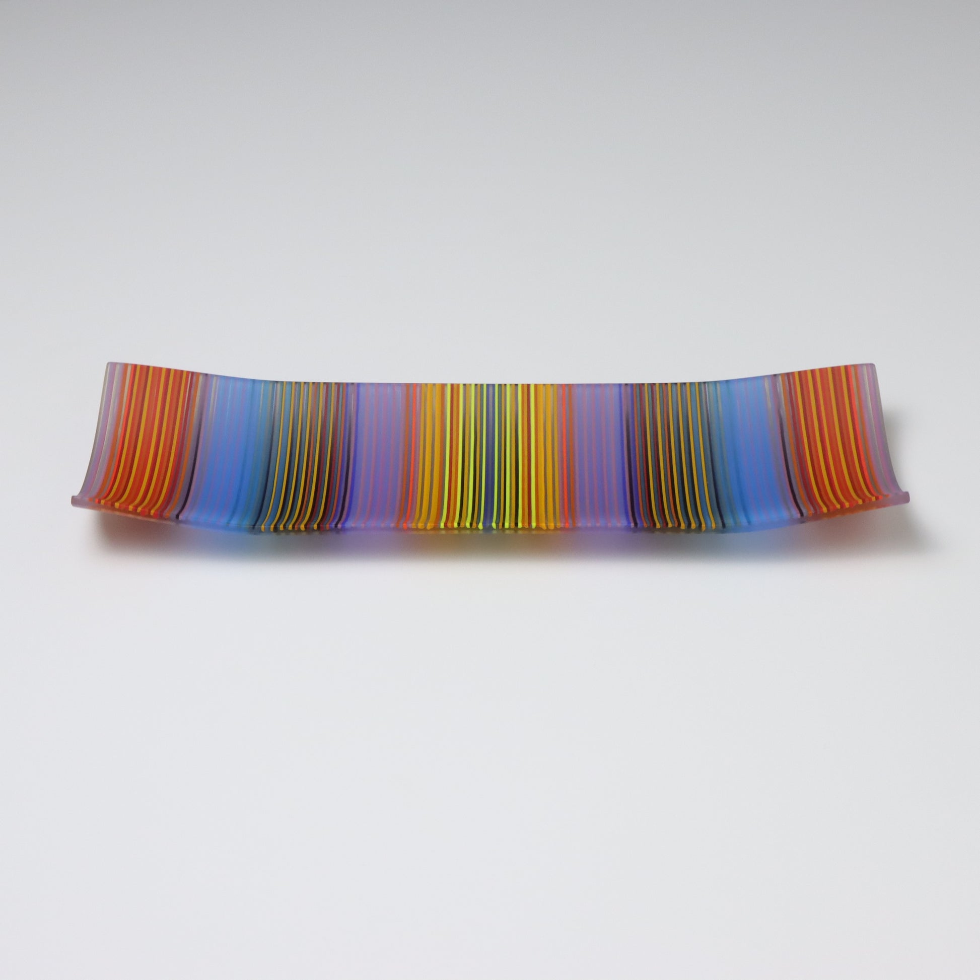 An exquisite ColourWave Glass plate with a rectangular shape, featuring a striking pattern of vertical stripes in a bold colour palette including shades of blue, purple, orange, yellow and green, with many more seen up close. The design is complemented by the plate’s corners, which gracefully curve upwards, giving it a refined and elevated appearance.