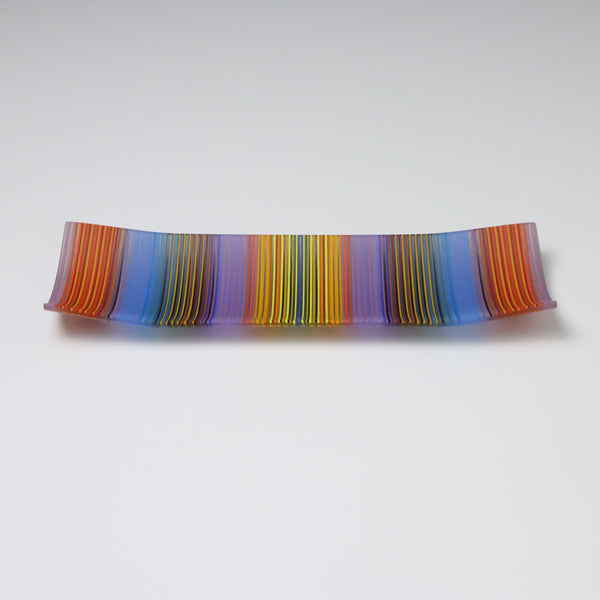 An exquisite ColourWave Glass plate with a rectangular shape, featuring a striking pattern of vertical stripes in a bold colour palette including shades of blue, purple, orange, yellow and green, with many more seen up close. The design is complemented by the plate’s corners, which gracefully curve upwards, giving it a refined and elevated appearance.