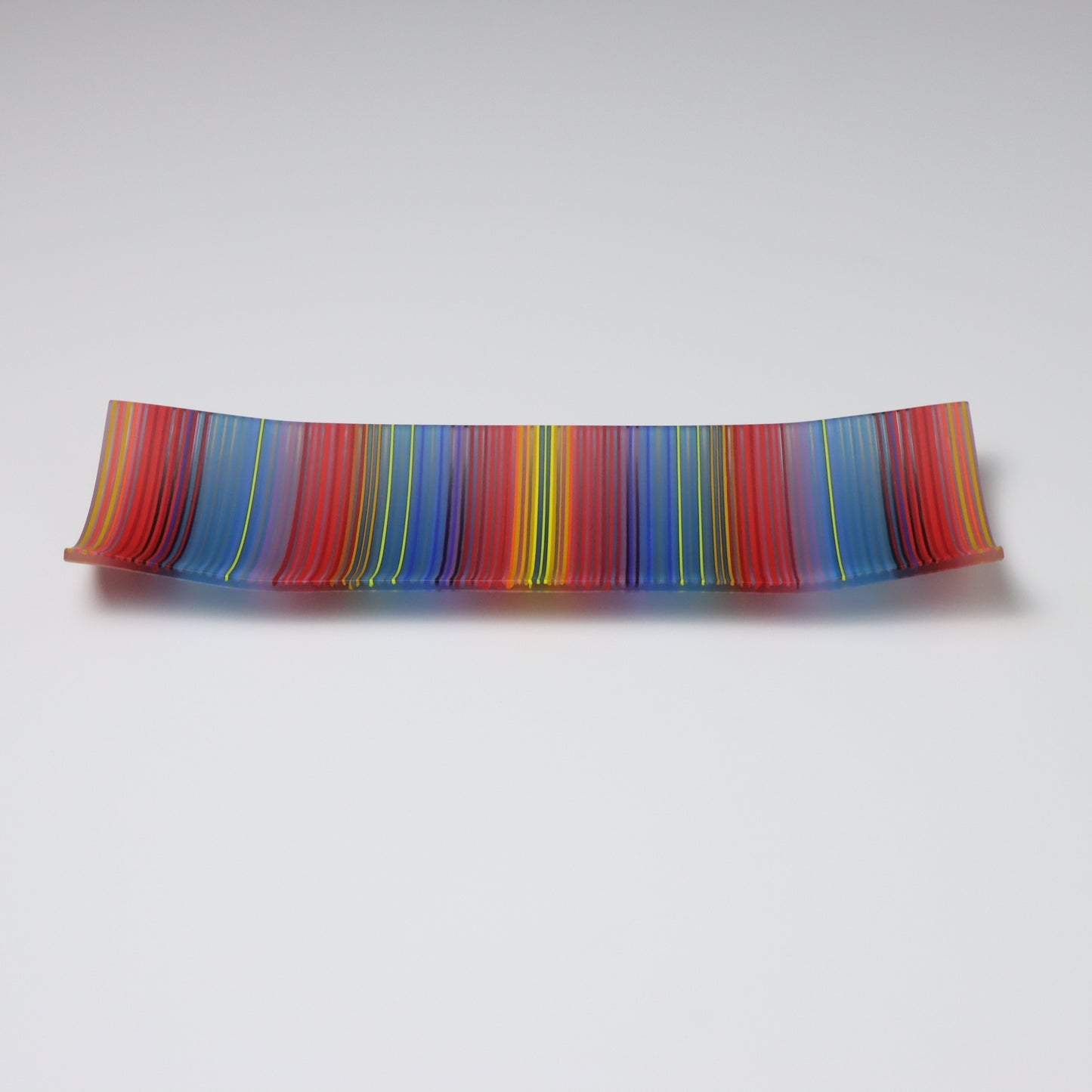 A ColourWave Glass fused glass plate, arrayed with vertical stripes in vibrant red, blue, yellow, and green. The plate’s rectangular shape is enhanced by the corners that curve upward, forming a shallow, dish-like appearance. Set against a grey background, the plate’s rich stripes and distinctive design are highlighted, making it an eye-catching piece.