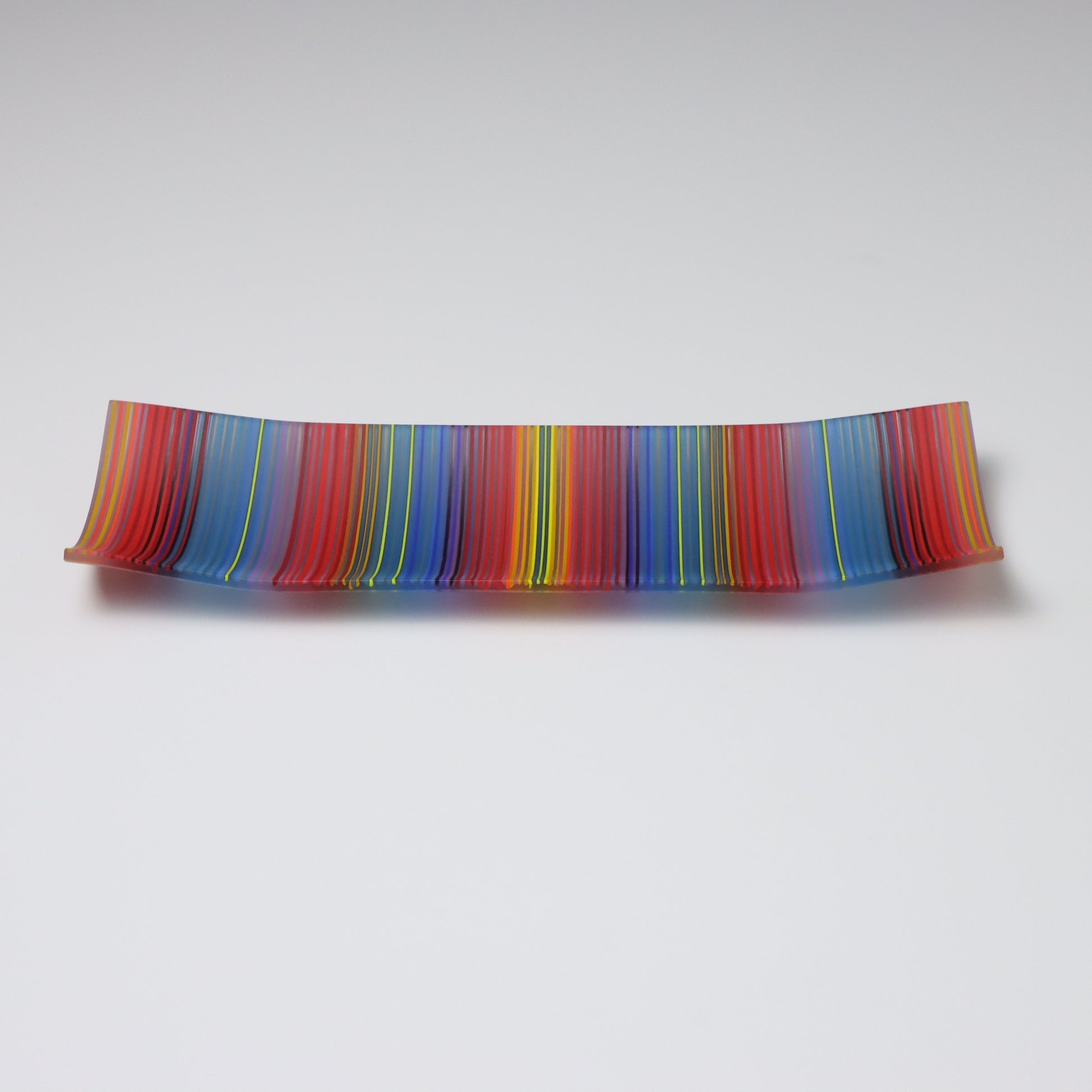 A ColourWave Glass fused glass plate, arrayed with vertical stripes in vibrant red, blue, yellow, and green. The plate’s rectangular shape is enhanced by the corners that curve upward, forming a shallow, dish-like appearance. Set against a grey background, the plate’s rich stripes and distinctive design are highlighted, making it an eye-catching piece.