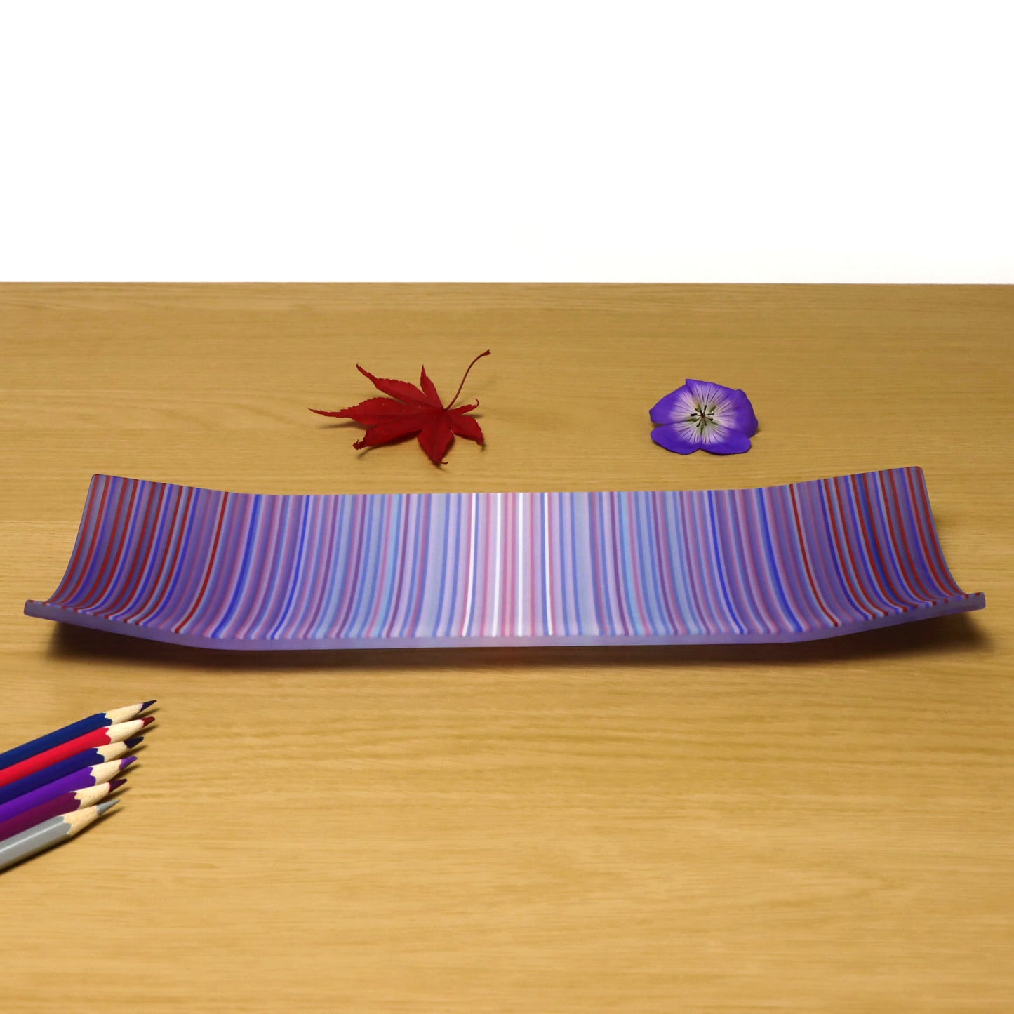 A striking ColourWave Glass fused glass plate. The design features a captivating blend of purple, blue, and subtle pink stripes. The plate is presented on a wooden surface, complemented by a red maple leaf and a purple flower to the right, and a selection of complimentary coloured pencils out in the left corner. This setup accentuates the plate’s bright colours and sleek, modern design, making it a captivating centrepiece for interior decoration