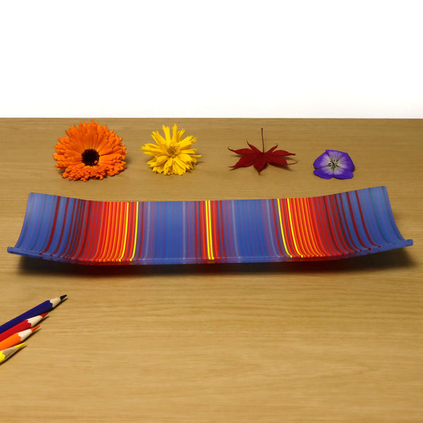 A ColourWave Glass fused glass plate featuring a harmonious blend of blue, red, and yellow stripes. The plate is set on a wooden surface, accompanied by a vivid orange gerbera, a bright yellow daisy, a red maple leaf, and a purple pansy. Coloured pencils in harmonising colours lay below the plate, complementing the striking colour palette and fluid design of the decorative fused glass.